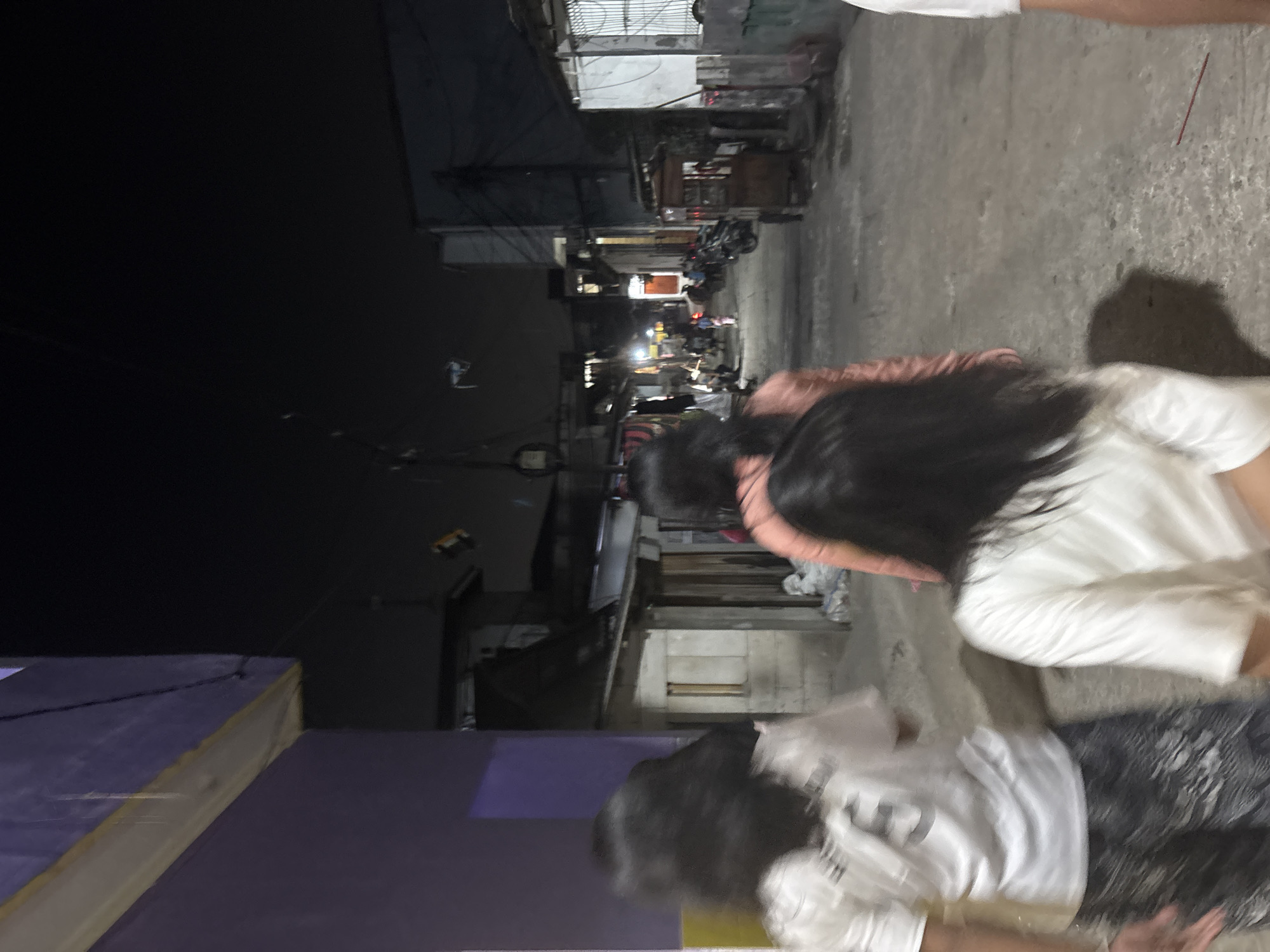 The image depicts a nighttime scene in a street, likely in North Jakarta, Indonesia, given the architectural style and the presence of what appear to be local businesses and residences. The foreground features three individuals, their faces blurred, walking away from the camera. The person on the left wears a white t-shirt with a number on it and patterned trousers, and is holding a white bag. The person in the middle wears a pink top, and the person on the right wears a white t-shirt.
The street is paved with concrete and shows some wear and tear. To the left, a purple building with a yellow trim is visible, partially obscuring the view of buildings behind it. To the right, there are various structures, including a small building with a corrugated metal roof and a food cart. Several motorcycles are parked along the side of the street. In the background, more buildings and the dark night sky are visible, with some faint lights indicating activity. The overall atmosphere is that of a residential or commercial street at night, with a sense of everyday life unfolding.