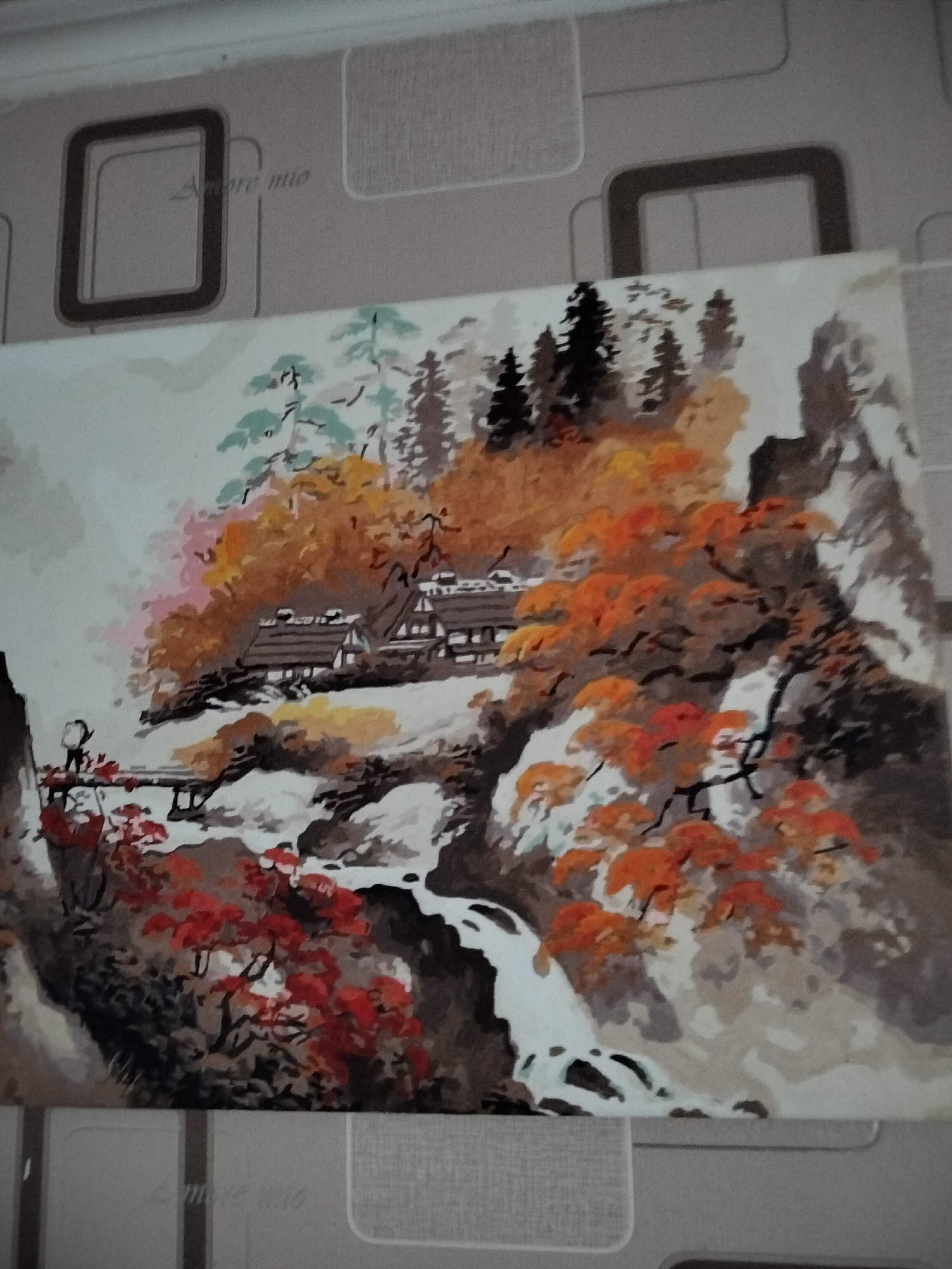 The image is a painting depicting a traditional Japanese landscape with mountainous terrain, a waterfall, and several buildings that appear to be houses with thatched roofs. Autumn foliage is prominent, with trees showcasing vibrant red, orange, and yellow leaves. A small wooden bridge spans a stream leading to the houses, and a solitary figure can be seen on the bridge, possibly an individual carrying a load. The background is a blend of misty grays and whites, suggesting a cloudy or foggy atmosphere. The style of the painting appears to be that of a traditional landscape artwork, possibly done in watercolor or gouache. The painting is placed against a background of wallpaper with a repeating pattern of squares and a text that says "Amore mio". There is no visible connection between the painting and the city of Nizhnedonskoy, Russia.