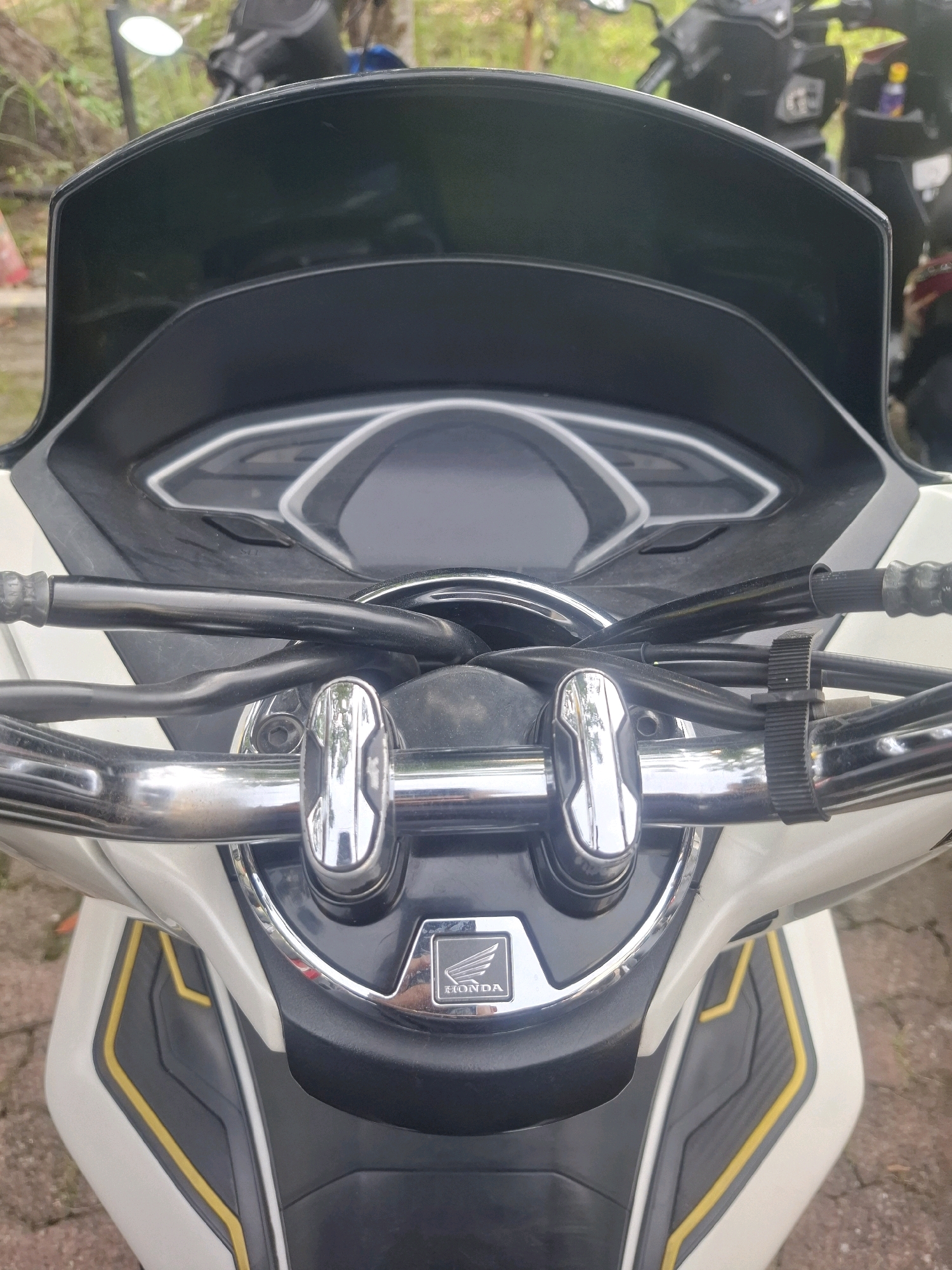 The image shows a close-up, head-on view of the handlebars and front fairing of a white and black scooter, likely in Medan Estate, Indonesia. The scooter features a dark windshield, chrome handlebars with black grips, and a prominent "Honda" logo on the front. The scooter is parked on a brick-paved surface, with hints of greenery and other scooters in the blurred background. No people are visible in the frame.