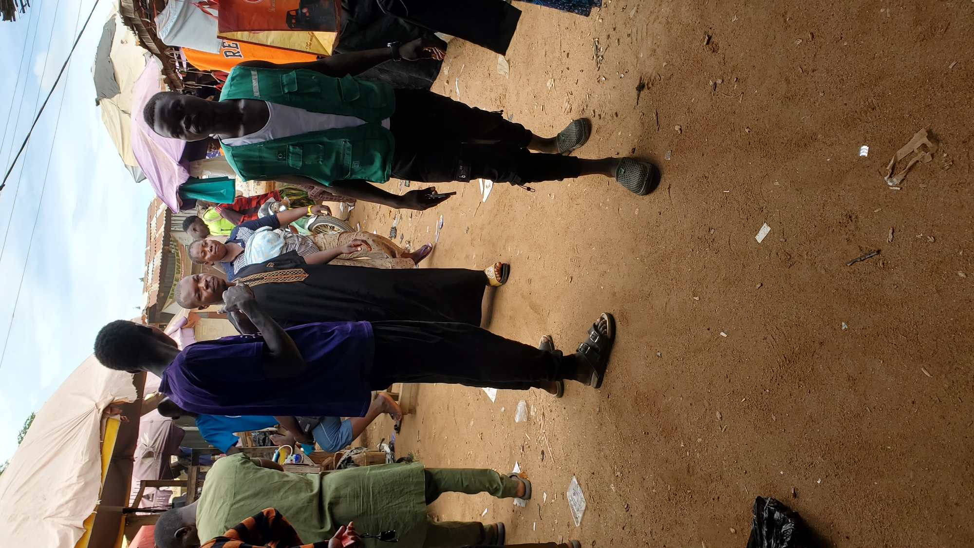 This is a street scene in Jalingo, Nigeria, depicting a market with several people present. In the foreground, a person wearing a green vest and dark pants walks towards the viewer, holding a mobile phone. To their left, another person in a purple shirt and black pants stands with their back to the camera, gesturing with their left hand. Further back, a man in a black traditional robe is visible, with a woman wearing a striped shirt and patterned skirt behind him. The ground is sandy and littered with debris. Tents and market stalls are set up in the background, suggesting a bustling commercial area.