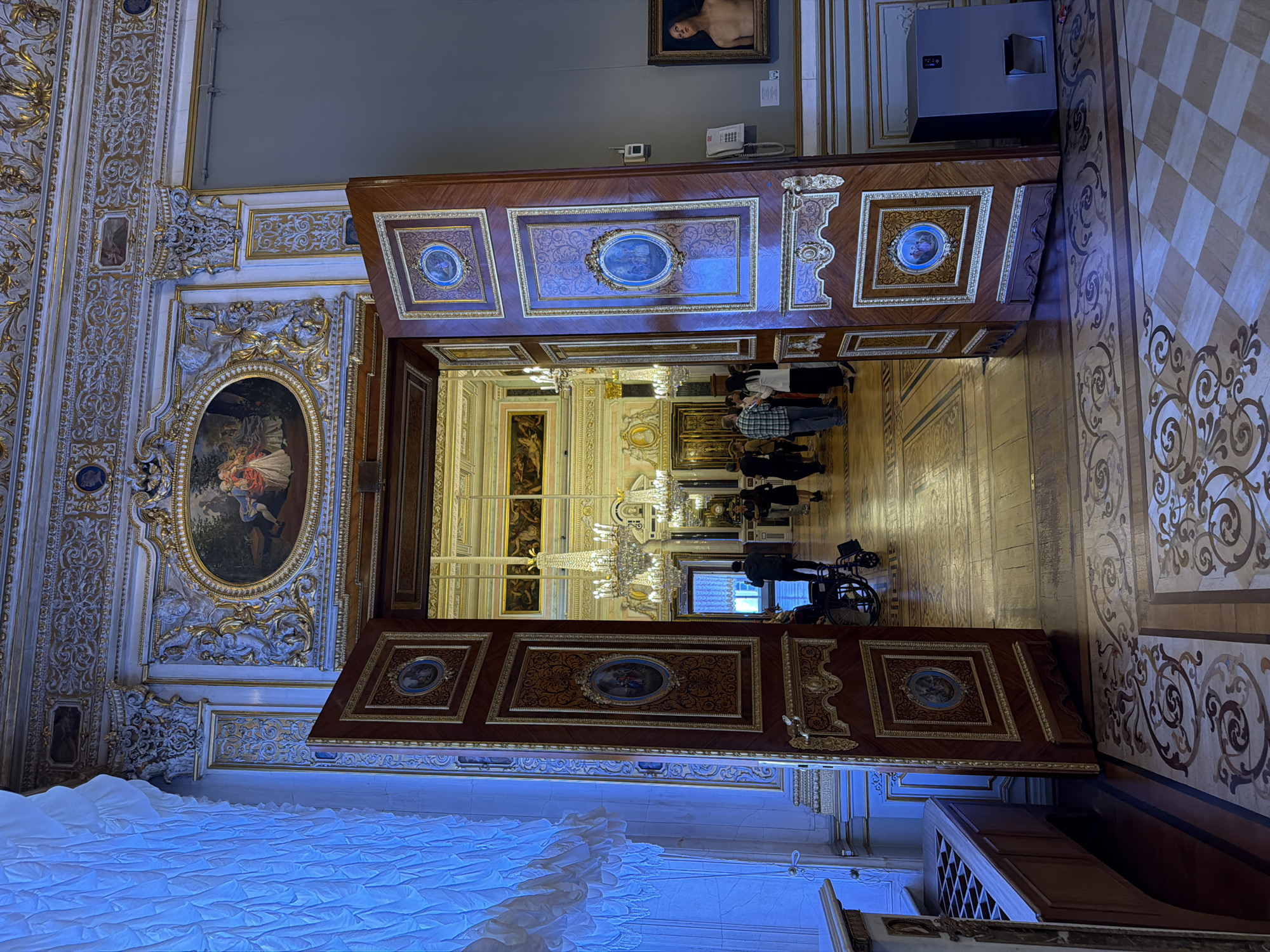 This media file depicts an interior scene within a grand, opulent building, likely a museum or palace in Saint Petersburg, Russia. The primary subjects are a group of people observing the surroundings, a set of large, ornate wooden double doors, and the richly decorated interior architecture.
The scene appears to be set during the daytime, judging by the artificial lighting that illuminates the space. The architecture is characterized by elaborate gold ornamentation, detailed moldings, large framed artworks on the walls, and a checkered floor pattern in one section. The double doors themselves are intricately decorated with inlaid wood and framed oval paintings, suggesting high craftsmanship and historical significance.
A group of people are visible, some standing and observing, while one individual is seated in a wheelchair. Their posture and focus suggest they are engaged in viewing the exhibits or appreciating the architecture.
Notable details include a large, ornate chandelier visible through the open doors, a contemporary grey appliance or structure near the ceiling on the right, and a piece of artwork depicting a nude figure hanging on a wall in the background. The floor also features intricate inlaid wood designs. There are no discernible signs of weather or specific time of day beyond artificial illumination. The overall impression is one of a well-preserved historical or cultural site, designed to impress and showcase artistry and grandeur.
