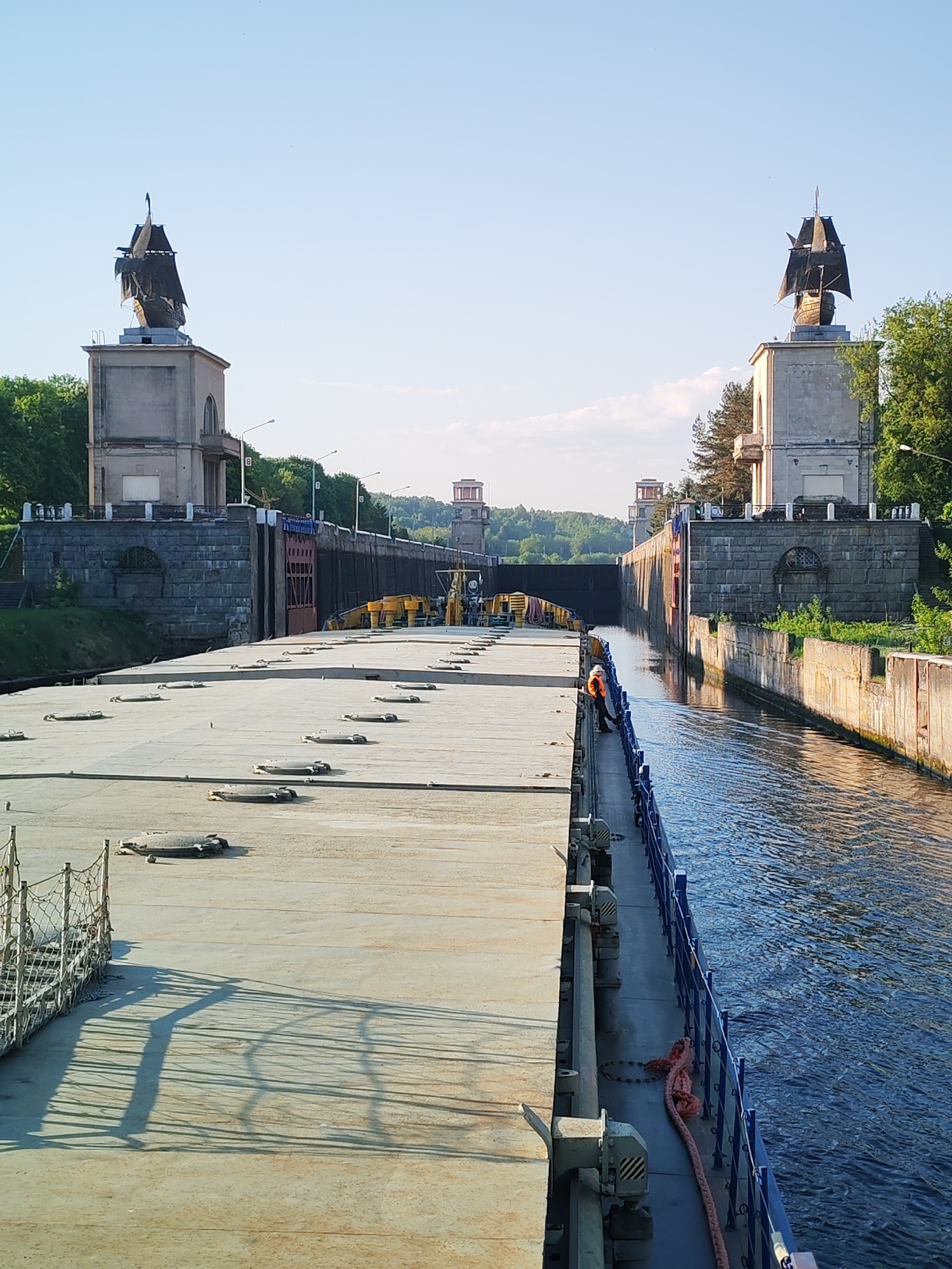 The image shows a view of the Moscow Canal in Yakhroma, Russia, specifically the lock structures. A large barge is visible in the foreground, taking up a significant portion of the frame. On the barge, a person wearing an orange safety vest is standing along the side. The canal is flanked by concrete embankments. On either side of the canal channel, architectural structures resembling towers are topped with sculptures of sailing ships. A bridge crosses over the canal in the distance, supported by smaller towers on either side. The sky is clear.
