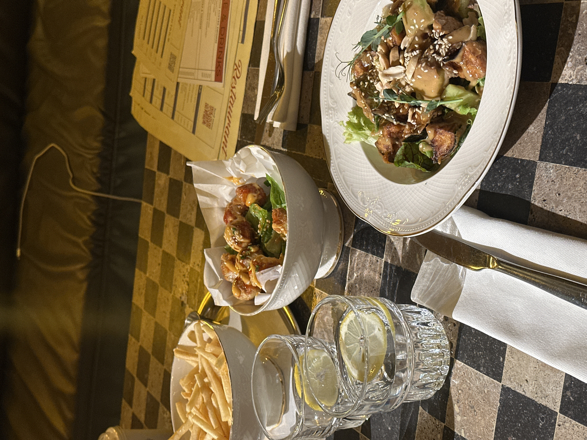 This close-up, top-down view captures a dining table set with various dishes and drinks, likely in a restaurant in Yerevan, Armenia. The table features a distinctive checkered pattern composed of alternating light brown and dark brown/black squares.
In the upper right portion of the frame, a white, ornate plate with a gold rim holds a main course. This dish appears to be a salad or a meat/tofu preparation, consisting of fried or roasted pieces coated in a light brown sauce, garnished with crushed peanuts and sesame seeds, and fresh microgreens or herbs over a bed of lettuce. To the left of this main plate, a smaller white bowl with a gold base contains similar fried pieces, also drizzled with sauce and green garnish. Further to the bottom left, a portion of a larger white bowl filled with golden-brown French fries is visible.
Two clear, textured drinking glasses with slices of lemon are placed towards the bottom center of the image, one appearing fuller than the other. A white cloth napkin with a gold-toned knife and a silver-toned fork rests neatly beside the main plate.
On the far left, several stacked yellowish papers, likely menus or bills, are visible. The word "Restaurant" is prominently displayed in an elegant script on one of the papers, with "DRINKS" also visible further down. Several QR codes are printed on these documents. A thin white cable runs across the dark surface to the left of the papers.
The lighting is dim and warm, suggesting an evening setting or a cozy indoor ambiance, with shadows cast by the plates and glasses on the tabletop. The overall scene depicts a prepared meal awaiting consumption.