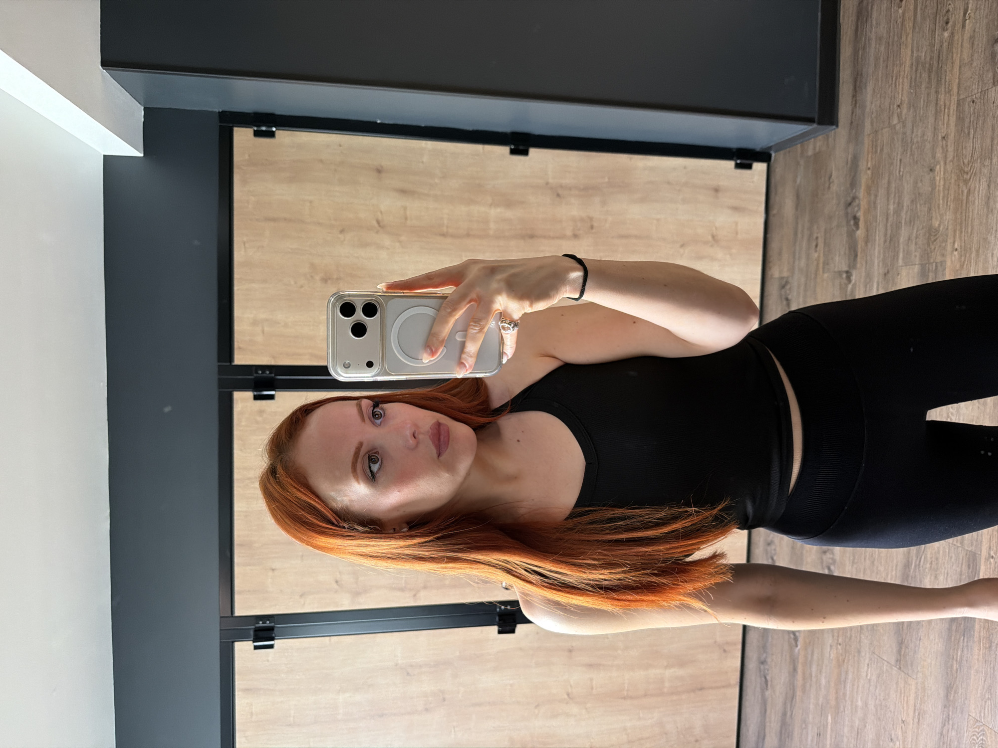 A fair-skinned, red-haired woman is taking a selfie in what appears to be an interior space. She is holding a phone in her right hand, and her left hand is holding the phone. She is wearing black workout attire, including a tank top and leggings. The background consists of a wall with wooden panels and dark frames. The location of this photo is not discernible as Paris, France, or any other location.