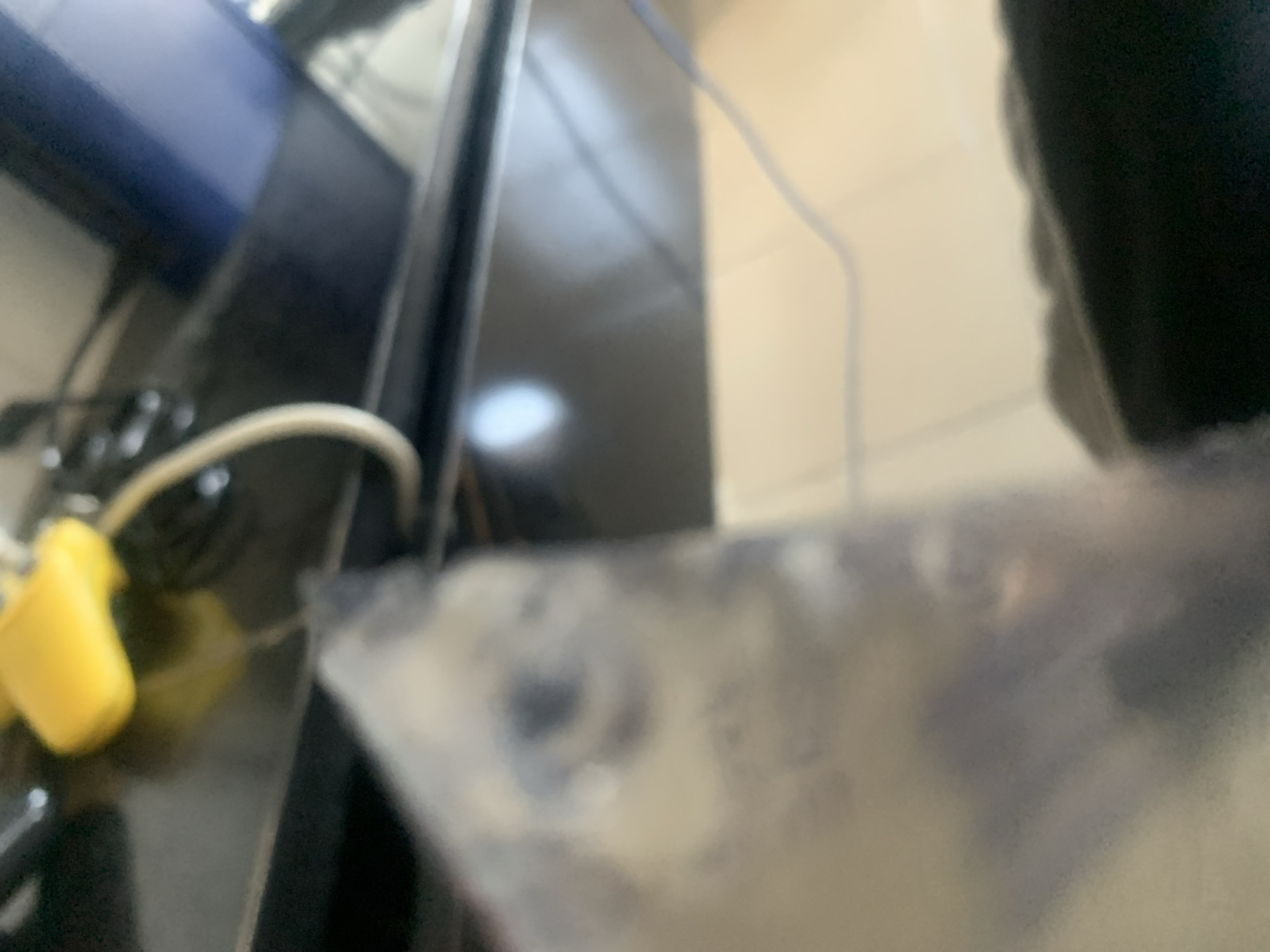 The image depicts a close-up, out-of-focus view of a cluttered surface, likely a desk or table, in Jalingo, Nigeria. In the foreground, a blurred, translucent object, possibly a plastic bag or container, partially obscures the scene. Within this object, a faint impression of a face or an object with distinct features, such as an eye and nose, is visible. The background reveals a dark, reflective surface, possibly a television screen or a glossy tabletop, with a yellow object and a white cable lying on it. Another cable, possibly blue, snakes across the reflective surface and extends out of frame. The overall environment appears to be an indoor setting, likely a residential or office space, with artificial lighting contributing to the reflections and blurriness. The image lacks any specific contextual clues to pinpoint an exact location within Jalingo.