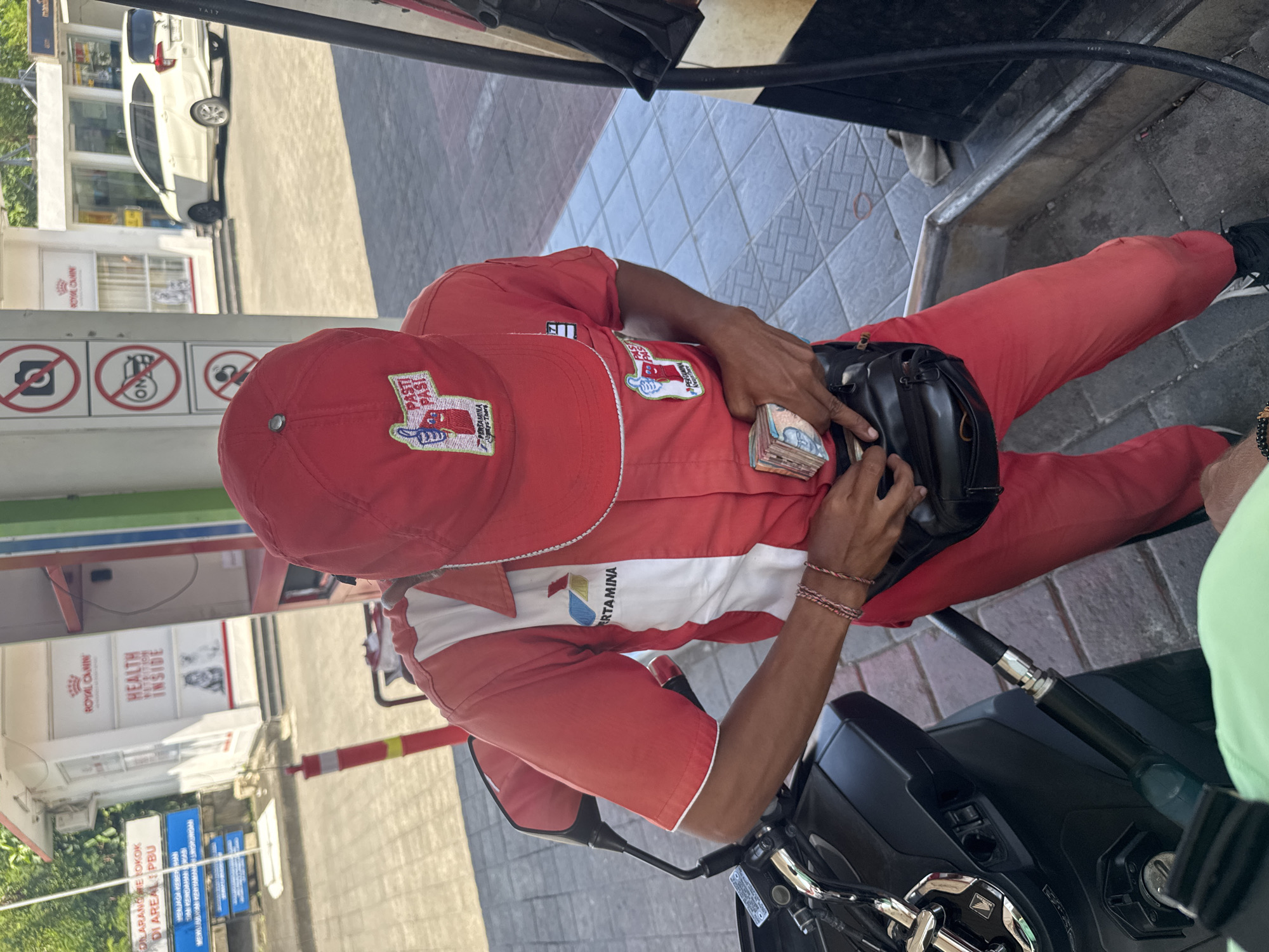 The scene depicts a Pertamina gas station in North Kuta, Indonesia. A gas station attendant, wearing a red uniform and cap with the Pertamina logo, is seated on a black motorcycle. He is counting a stack of Indonesian Rupiah banknotes while near the gas pump. Signage in the background indicates no smoking and the location of Pertamina gas station, along with storefronts of Royal Canin.