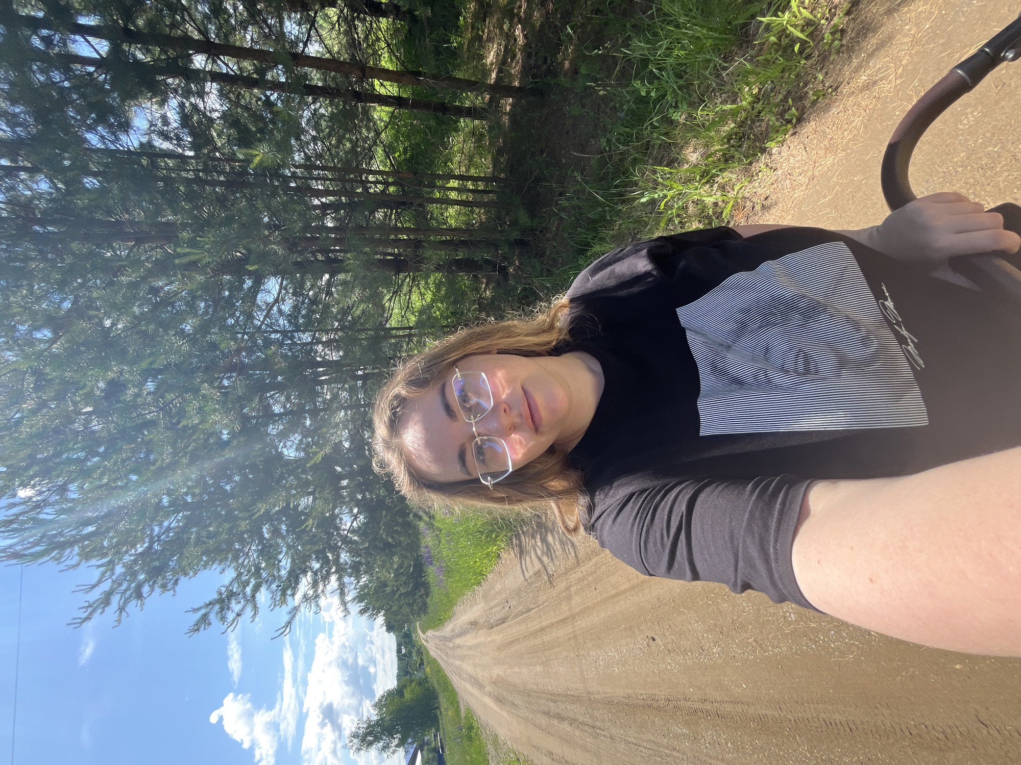A fair-skinned woman, facing the camera and appearing to take a selfie, is positioned on the left side of a dirt road in a rural or semi-rural setting in Сосново, Russia. She has light brown hair, wears clear-framed, slightly oversized glasses with a subtle pink lens tint, and a black t-shirt featuring a white graphic design of varying horizontal lines and faint text. Her left hand is visible gripping a dark brown, padded handle, consistent with a stroller. The dirt road, marked by tire tracks, recedes into the distance under a bright blue sky with scattered white cumulus clouds. To the immediate left of the road, green grass and purple flowers are visible, with distant houses and trees under the sky. To the right of the road, a dense line of pine trees stands, through which sunlight prominently streams. The ground beneath the trees is covered with grass, dirt, and pine needles. The scene is illuminated by strong sunlight, indicating a clear day.