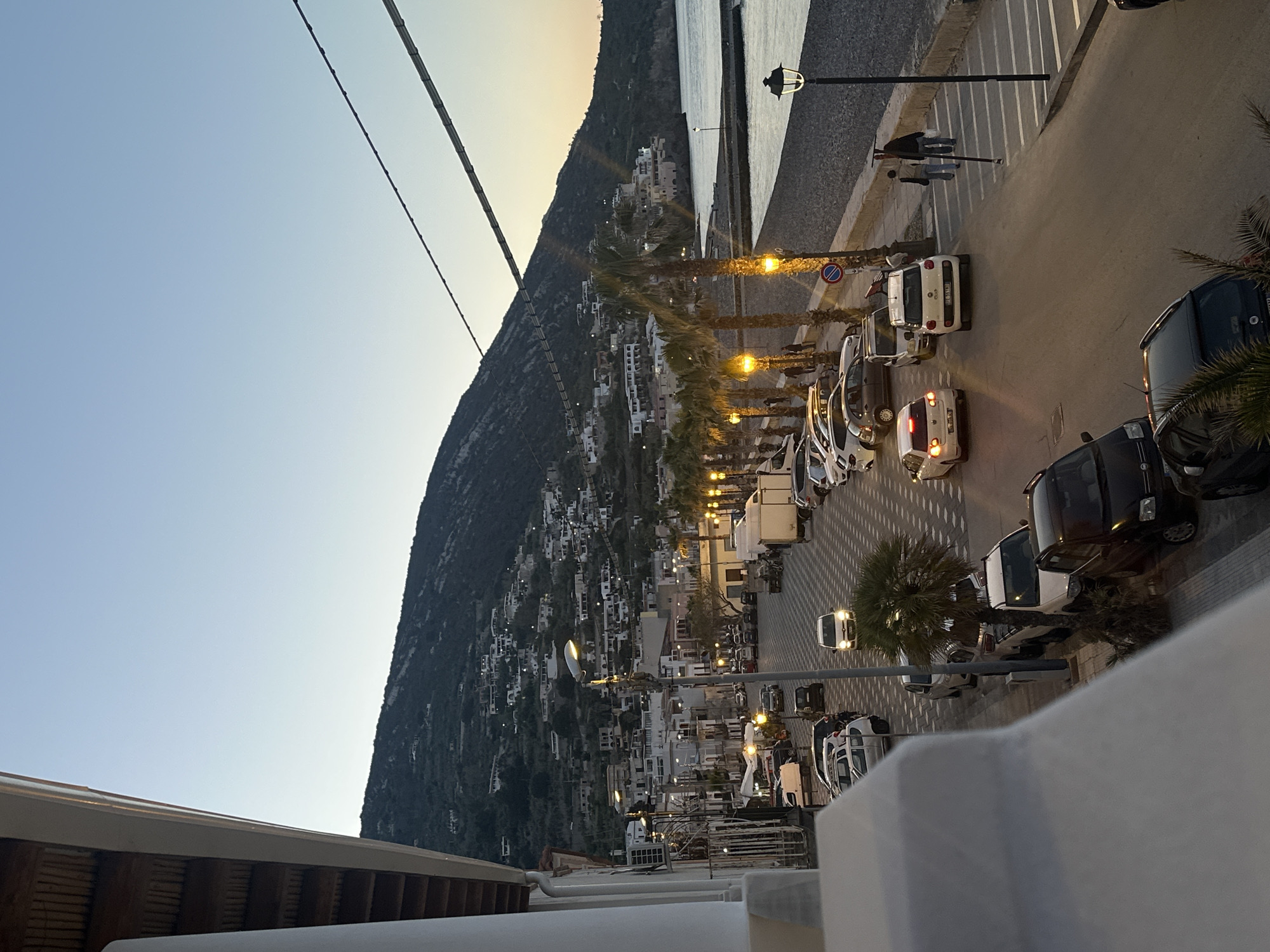 View from an elevated vantage point looking along a street lined with parked cars, palm trees, and streetlights, bordered by a beach and a steep, densely built-up hillside at dusk in Lipari, Italy. Three people are visible walking along the beach promenade.