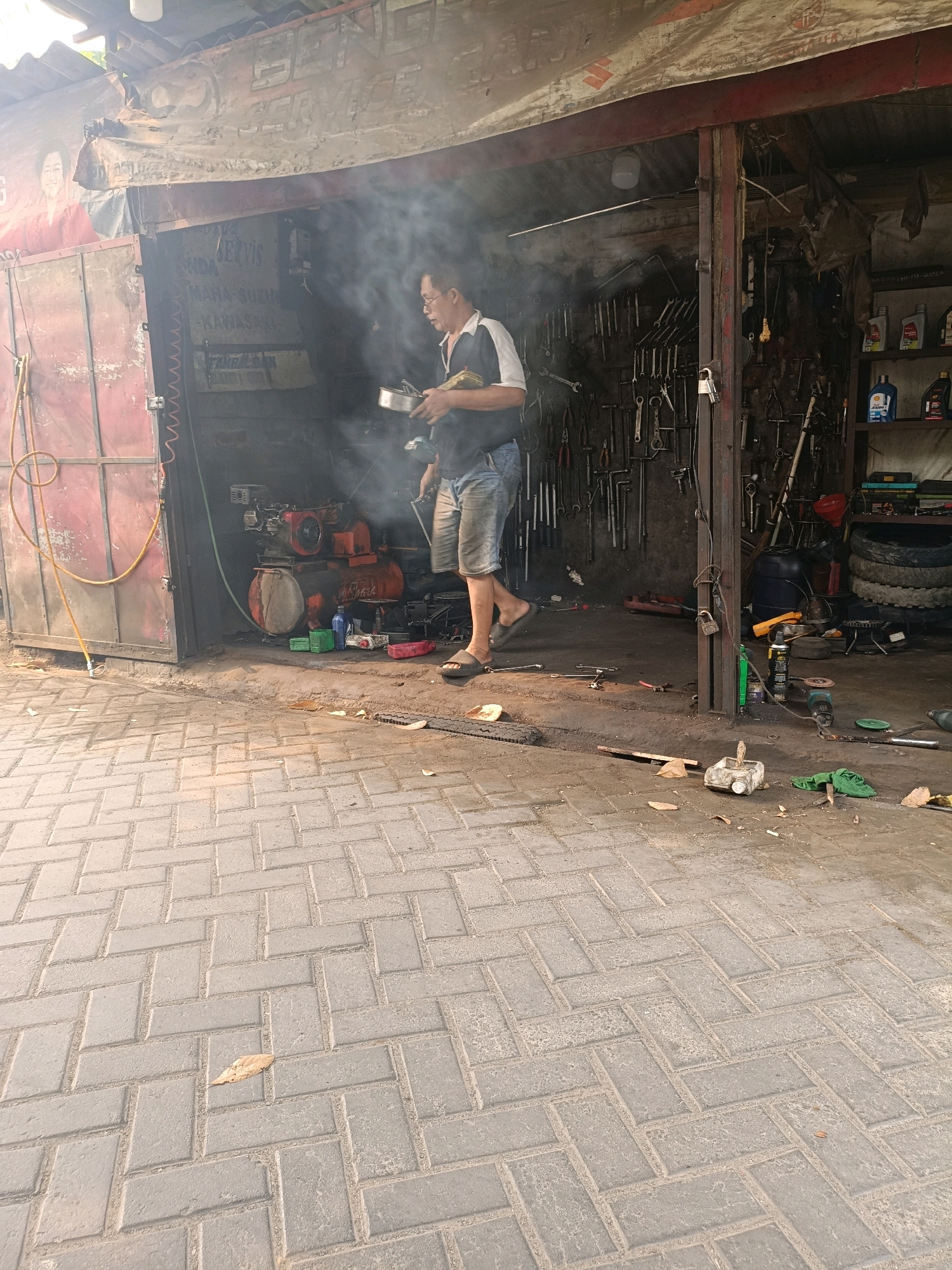 The image shows a man working in a workshop. The workshop appears to be an open-air garage with a corrugated metal roof. The man is wearing a dark blue shirt and denim shorts, and he is holding a metal bowl. There is a lot of smoke in the air, indicating some kind of activity is taking place. The workshop is filled with tools and equipment, including an air compressor, tires, and various hand tools hanging on the wall. The floor is made of interlocking pavers. The background shows a red metal door with a yellow hose attached to it. On the wall of the workshop, there are some posters, one of which depicts a woman. The overall scene suggests a busy and functional repair shop. The presence of the poster with the woman and the specific style of the workshop point to a location in Tangerang, Indonesia.