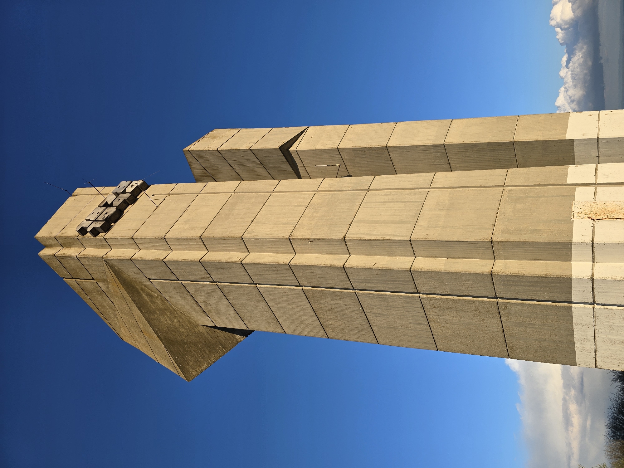 The image shows a view of the Buzludzha Monument in Pobeda, Bulgaria, from below, angled towards the sky. The concrete structure is composed of stacked, rectangular modules. The monument has a sloping, triangular base section. A series of protruding, cube-like elements are located near the top of the structure, as well as antenna wires. The background is a clear blue sky with clouds and landscape elements visible near the horizon.