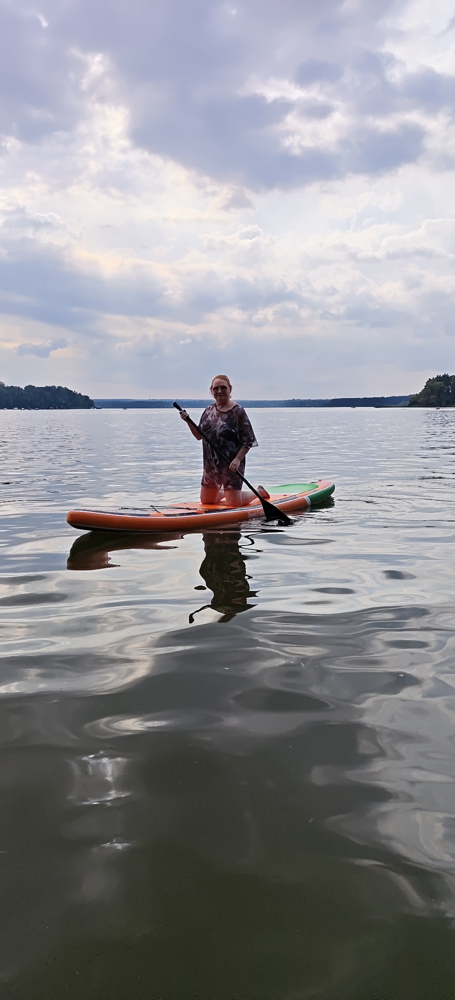 A woman kneels on an orange and green paddleboard in the middle of a calm body of water, holding a black paddle. She is wearing a camouflage-patterned dress and sunglasses. The water reflects the sky and the paddleboard, creating a rippling effect. In the background, a distant treeline borders the water, and a cloudy sky hangs overhead. The location is identified as Пятница, Russia, though no specific city landmarks are visible in the image.
