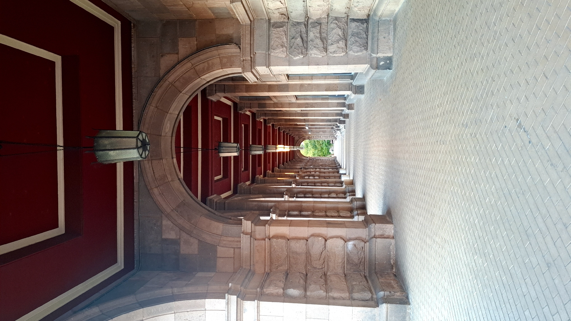 The image depicts a long, covered walkway with an arched entrance and a repeating series of arches supported by stone columns. The walkway has a patterned brick floor and a red ceiling with white trim. Several old-fashioned lanterns hang from the ceiling. In the distance, the walkway opens to an area with green trees and foliage. The architecture suggests a historical building or public space. This location is the South Entrance of the National Palace of Culture (NDK) in Sofia, Bulgaria. There are no people visible in the image.