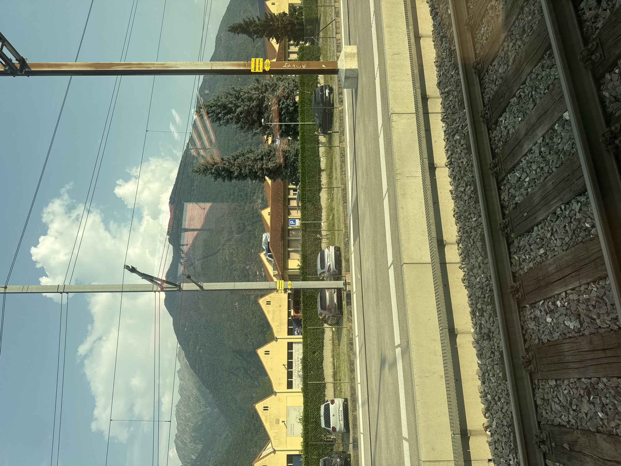 The image depicts a view from a train window, looking out at a railway track in the foreground. Beyond the track, a concrete platform is visible, followed by a fenced-off area with several parked cars and a row of yellow industrial buildings with sawtooth roofs. Tall evergreen trees are scattered in front of the buildings. In the background, a large, tree-covered mountain range rises under a partly cloudy sky. A tall, rusty metal pole with electrical wires attached stands prominently on the right side of the frame, and a yellow sign with Italian text is mounted on it. The scene appears to be in Bellinzona, Switzerland, given the signage and mountainous landscape characteristic of the Ticino region. There are no people visible in the image.