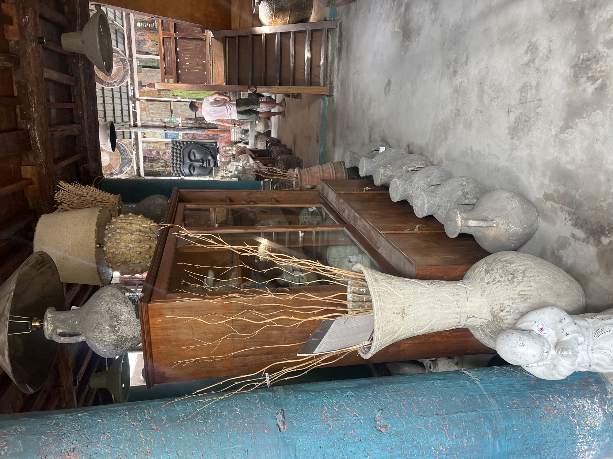 The image depicts an indoor scene in what appears to be an antique or handicraft shop in North Kuta, Indonesia. The shop features a rustic, eclectic decor with wooden beams and structures. In the foreground, a large, textured, pale gray vase with dried branches is prominent. To its left, a blue-painted pillar stands next to a wooden cabinet. A small, white statue of a person in prayer is situated in front of the large vase. A row of weathered, gray amphora-like vases are lined up on the floor extending into the background. Further in the background, a man wearing a light pink t-shirt and olive green shorts is walking away from the camera. He is near a wooden staircase leading to an upper level. Various decorative items, including a large black Buddha head sculpture and other ancient-looking artifacts, are visible throughout the space. The floor is a rough concrete surface. The overall lighting suggests an ambient indoor environment, possibly with natural light filtering in from unseen sources.