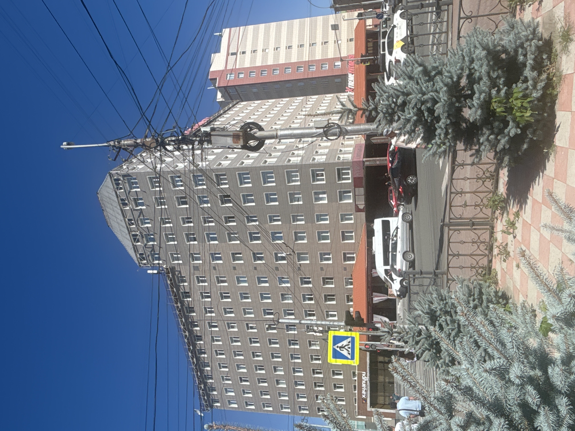 The image shows a view of a street in Novosibirsk, Russia, with a prominent building on the left. The building is tall and gray, with many windows. A pedestrian crossing sign is visible in front of the building. To the right, another building is partially visible, taller and lighter in color, with red and white vertical stripes. Power lines are strung across the sky, supported by a utility pole with many wires. In the foreground, there are evergreen trees and a sidewalk. Several cars are visible on the street, including a white van, a silver sedan, and a black car. A few people are walking on the sidewalk and crossing the street. The sky is clear and blue.