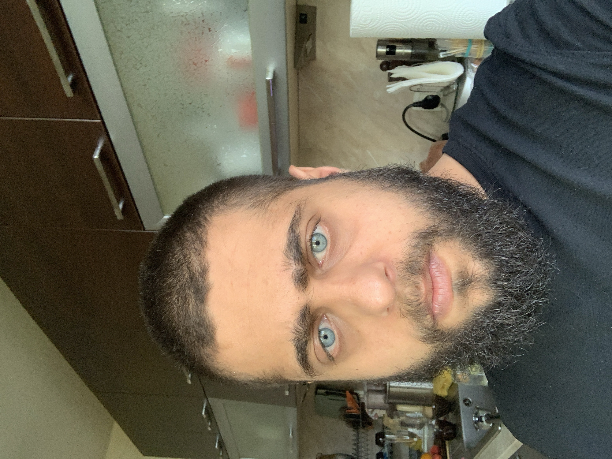 A rotated close-up portrait captures a man with striking light blue eyes and a full, dark beard with some visible gray strands. His short, dark hair appears buzzed on the sides. He wears a black t-shirt or hoodie and maintains a neutral expression, looking directly at the camera.
The background indicates an indoor setting, likely a kitchen or utility area. To the man's left, dark brown wooden cabinets with horizontal silver handles are visible, alongside a frosted glass panel. To his right, a roll of white paper towels rests on a holder above a light-colored counter. Various kitchen items, including a metal appliance, white napkins, and a black electrical cord plugged into an outlet, are scattered on the counter. The scene is illuminated by what appears to be artificial indoor lighting. No visible text is present in the image.