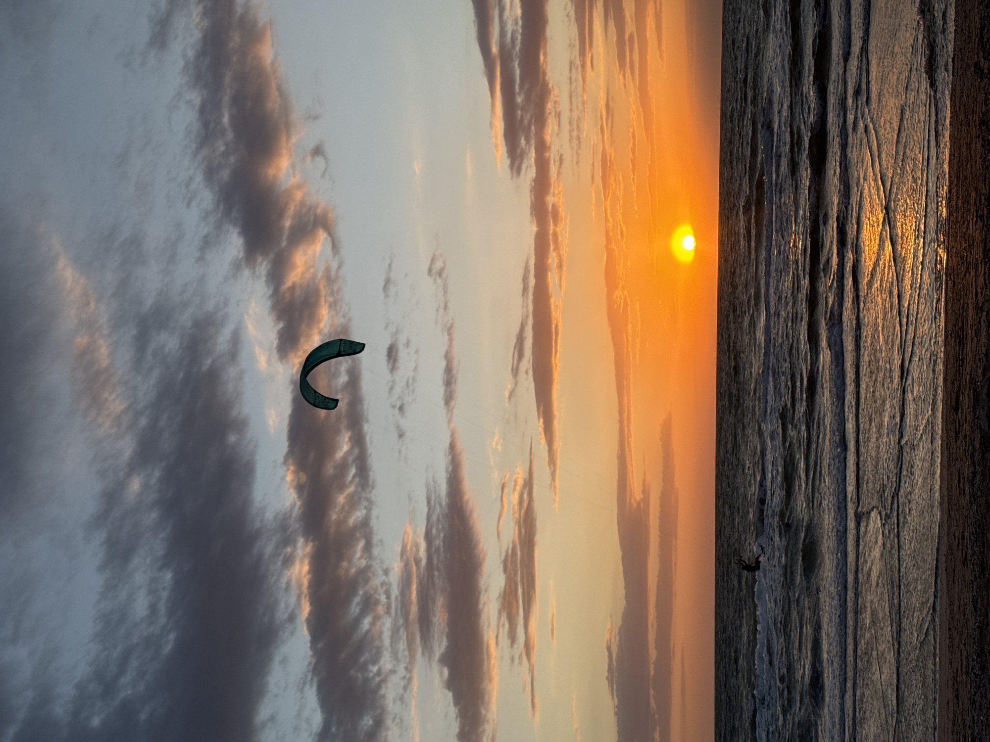 A vertical panoramic view of a sunset over water, possibly the Sea of Azov, near городское поселение Щелкино, Ukraine. The sky is a mix of dark grey clouds and orange sky. There is a kite surfer near the upper left corner. The sun is setting on the horizon on the right. The water is dark and reflective. There is a lone figure on the water near the bottom of the image.