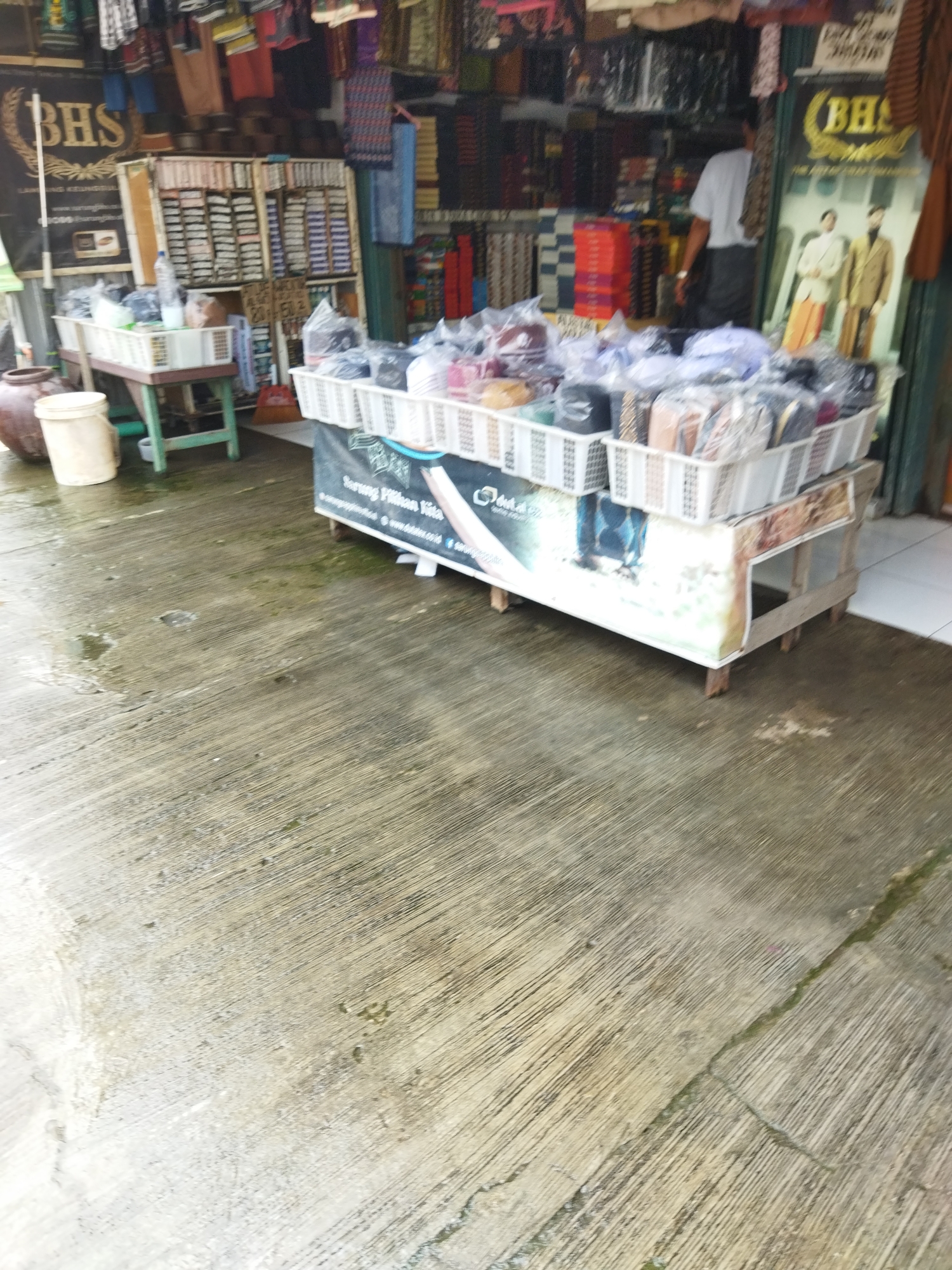 The image captures a street view of a shop in Suka Sari, Indonesia, possibly selling textiles and clothing. In the foreground, a wet concrete ground is visible with a few small puddles. A wooden cart with white plastic baskets filled with various items, likely clothing or fabrics, is positioned in the middle ground. A large dark brown ceramic pot and a light-colored plastic bucket are situated to the left of the cart. Behind the cart, a rack of shelves displays numerous items, possibly sarongs or similar textiles. The shop's facade features signage, including a large "BHS" logo, and displays of clothing, including two mannequins dressed in traditional attire on the right. A person wearing a white shirt and black pants is partially visible inside the shop. The overall scene suggests a commercial area with goods displayed outdoors.