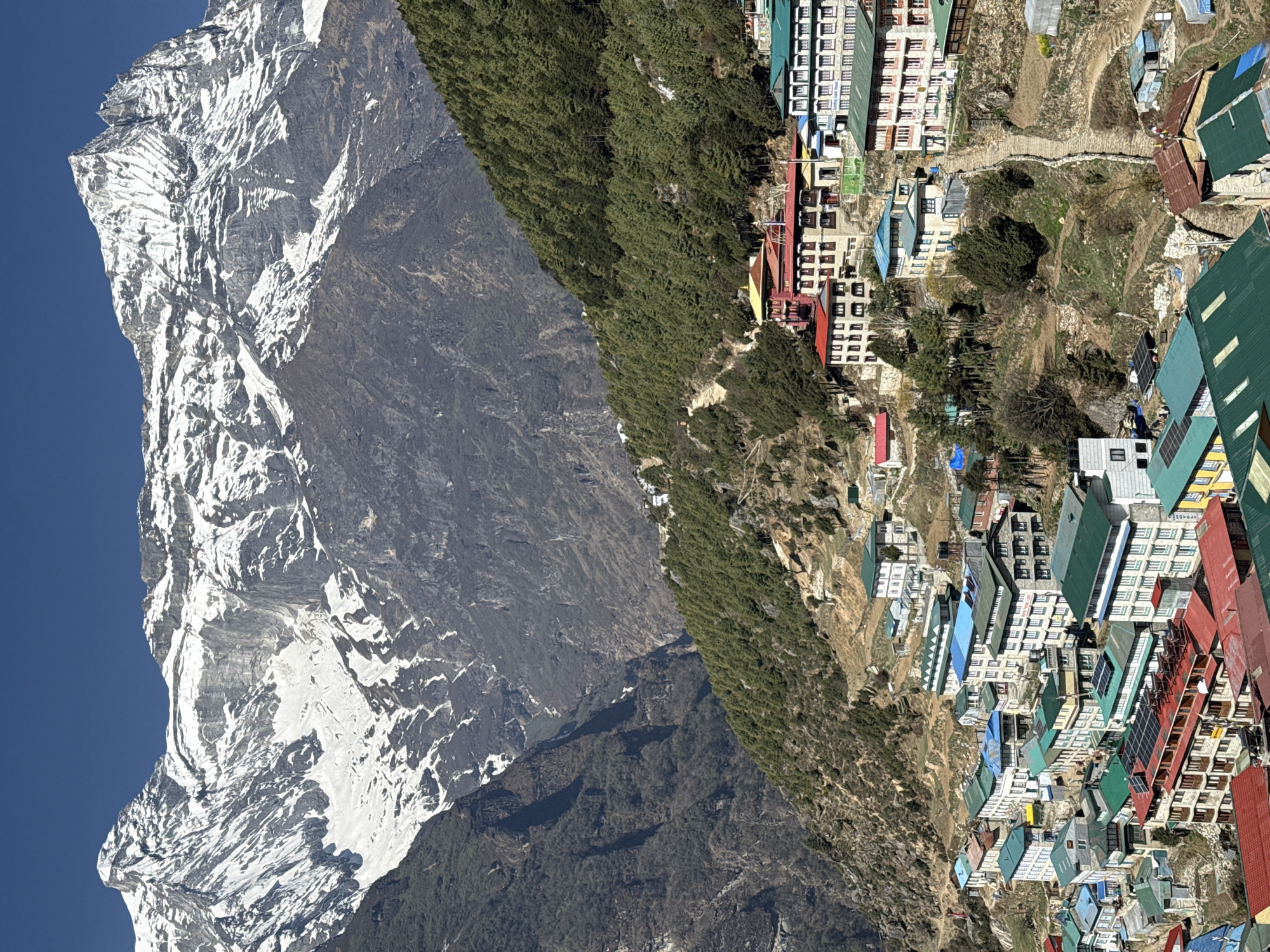 The image shows the town of Namche Bazaar in Nepal, nestled against a mountain range with snow-capped peaks under a blue sky. The town is built on a slope, with densely packed buildings featuring roofs of various colors, including green, red, and blue. A trail winds through the town and up the mountain. The mountain face is rocky with patches of snow and is partly covered with sparse vegetation. Trees line the edge of the settlement. The image captures the juxtaposition of the built environment and the natural landscape.