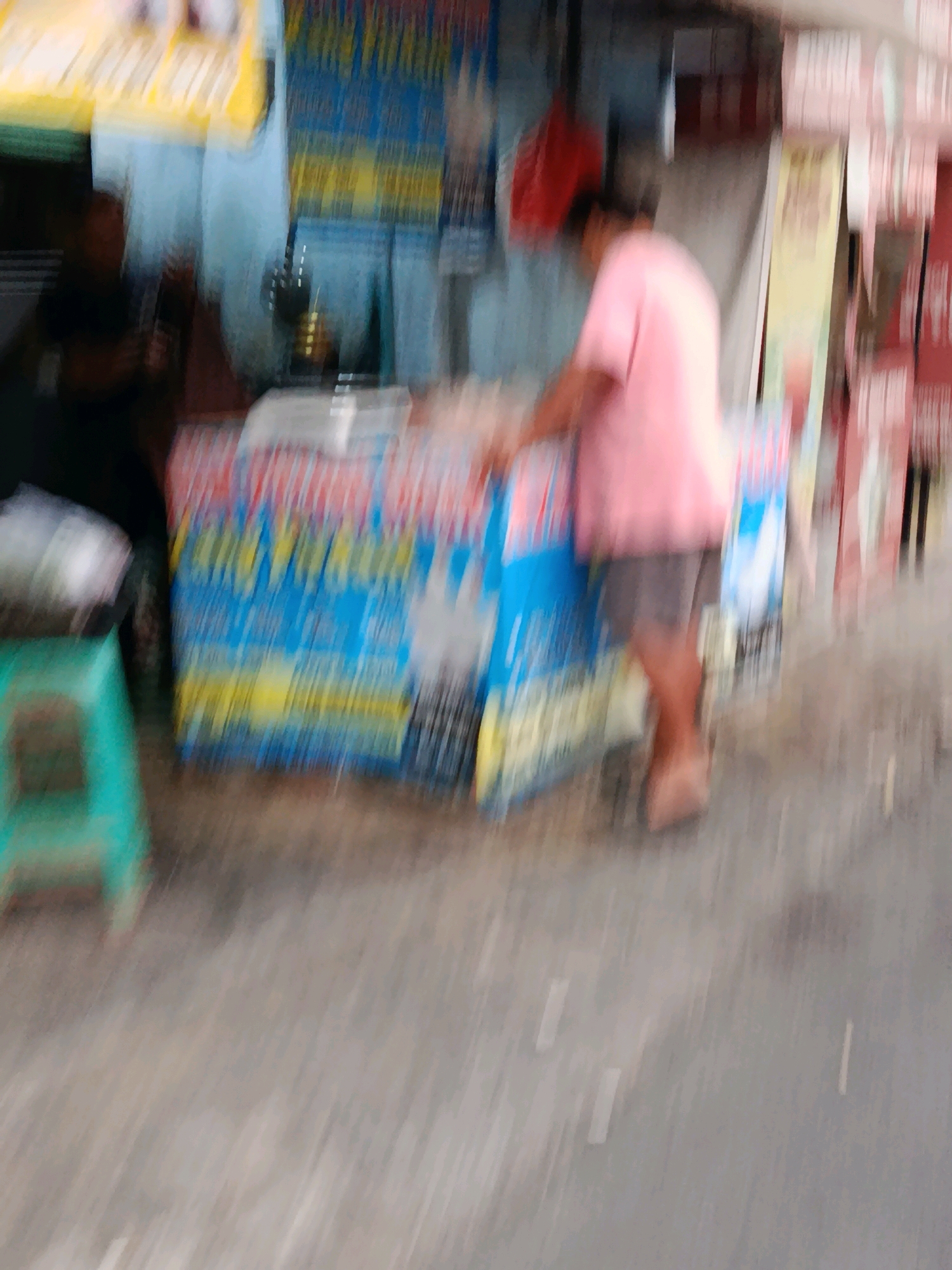 The image depicts an outdoor scene, likely a street-level market or food stall, in Bekasi, Indonesia. A person, appearing to be of average build and wearing a pink shirt and dark shorts, is visible from the waist down, standing behind a stall. The stall features blue and yellow signage with text that is largely illegible due to motion blur. To the left of the stall, a bright teal-colored stool is present. The ground appears to be wet or reflective, suggesting recent rain or a generally damp environment. The overall image suffers from significant motion blur, making detailed identification of specific items and individuals challenging. The background is indistinct, but hints of other structures and signs are present, consistent with an urban or semi-urban commercial area.