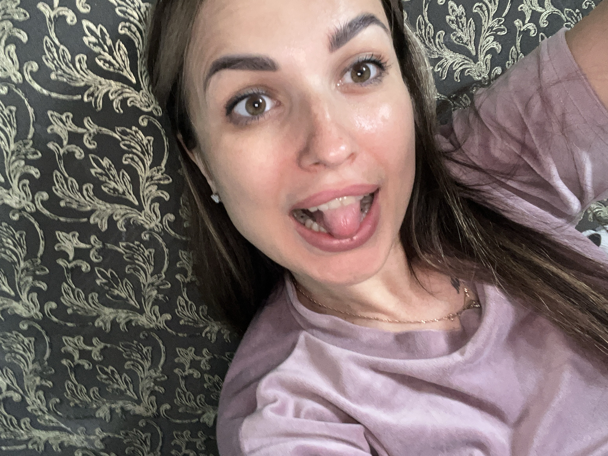 A close-up, high-angle, waist-up shot shows a woman lying on her back, looking directly at the camera with her mouth open and tongue sticking out slightly. She has brown eyes, dark eyebrows, and light brown hair. She is wearing a light pink, collared t-shirt and a delicate gold chain necklace. The background is a dark fabric with a raised gold floral pattern, possibly upholstery or wallpaper. The lighting is soft, highlighting her skin and the texture of the fabric.