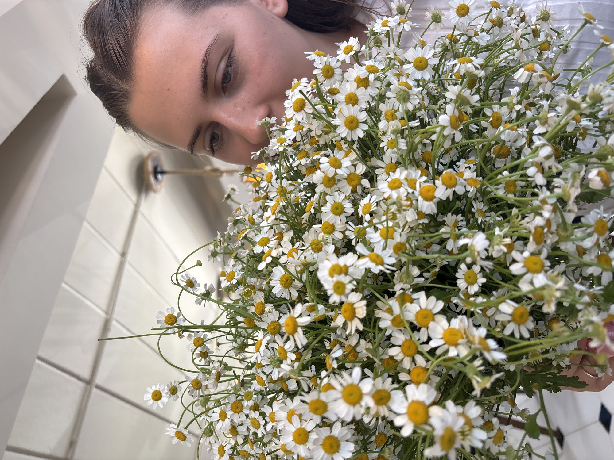 A woman is close to a bouquet of chamomile flowers with a yellow center and white petals. The woman's face is turned towards the flowers as if she might be smelling them. She has fair skin, brown hair, and is wearing a white shirt. The background includes a white wall and tile. The location is Phuket City Municipality, Thailand.