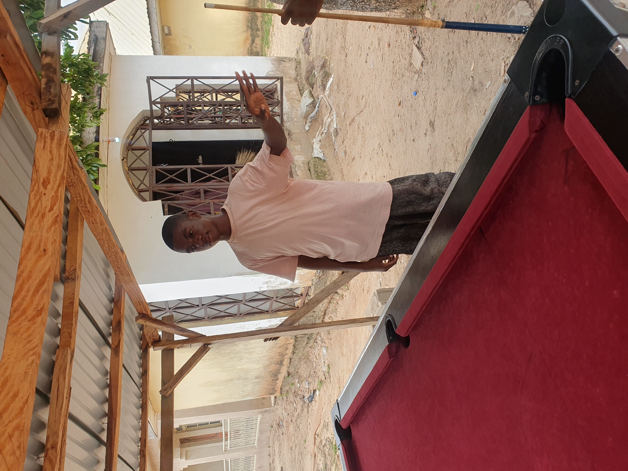 A young Black male stands under a wooden shelter with a corrugated metal roof, gesturing with his right hand. He is wearing a light pink t-shirt and dark patterned shorts. In the foreground, a red billiard table is visible. The ground is sandy, with scattered debris and some vegetation. A building with windows covered by metal bars is in the background, suggesting a location in Lokoja, Nigeria. A billiard cue is held vertically to the right of the frame.