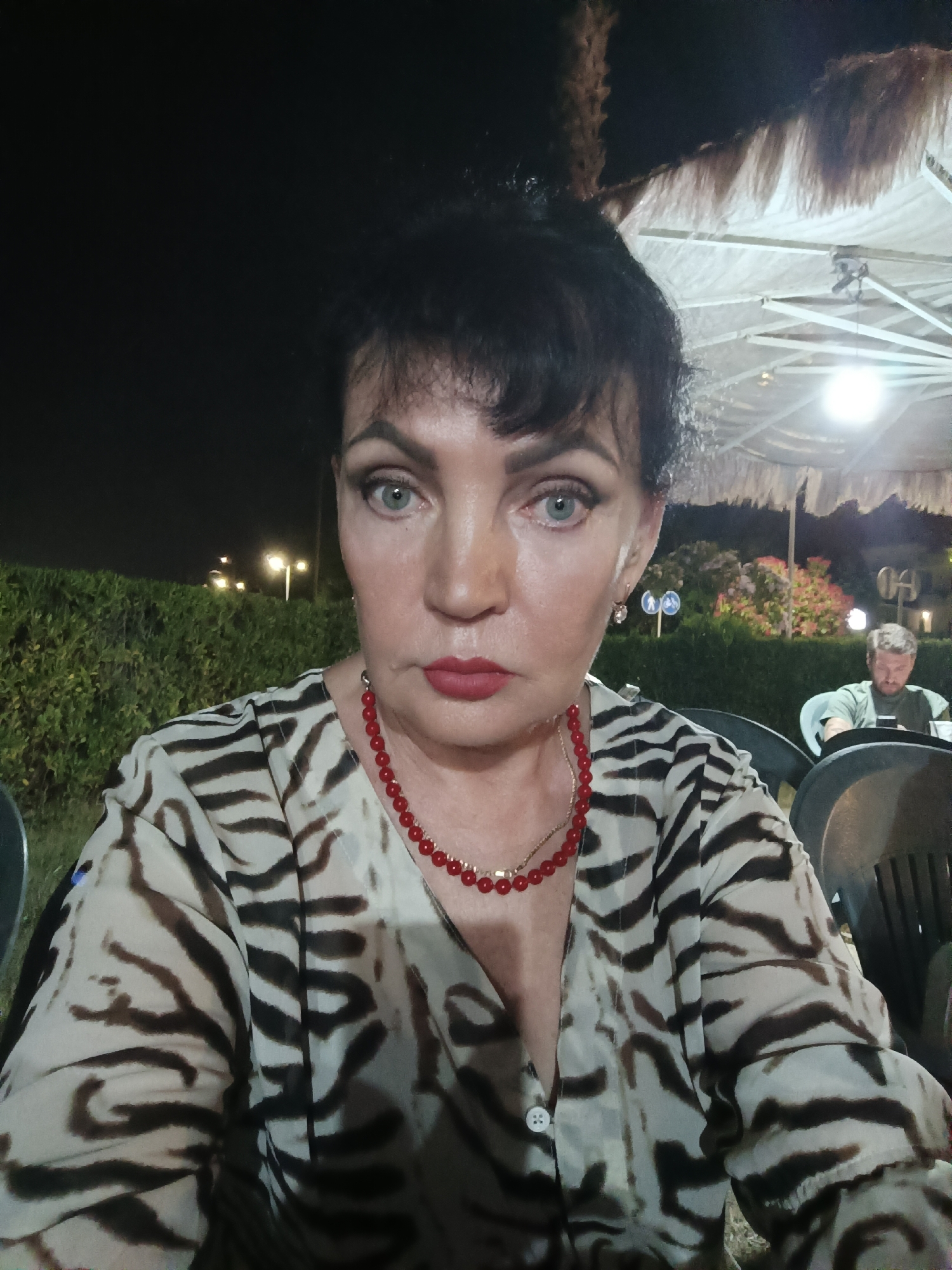 A woman with dark hair pulled back and bangs frames her face. She wears a zebra-print blouse and a red beaded necklace. Her makeup includes dark eyeliner and red lipstick. Her eyes are open and directed towards the camera. In the background, a man with light hair and a green shirt is seated at a table, looking down at his phone. He is blurry and out of focus. The setting appears to be an outdoor dining area at night, possibly in Giulianova, Italy, indicated by the presence of palm trees and architectural elements suggestive of a seaside town. A thatched-roof umbrella provides overhead cover. Streetlights illuminate the background foliage.