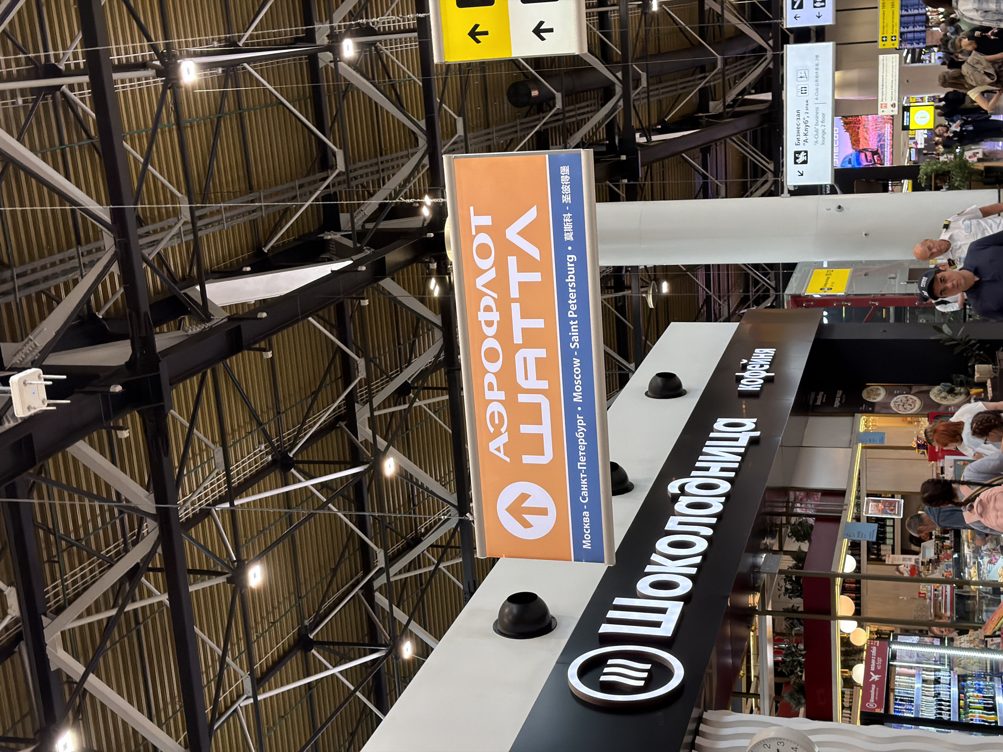 The image appears to be taken inside an airport terminal in Perepechino, Russia. The focal point is a prominent orange and blue sign with Russian text and an upward-pointing arrow, indicating Aeroflot Shatra airline. Below this, text reads "Москва - Санкт-Петербург - Moscow - Saint Petersburg - 莫斯科 - 圣彼得堡", suggesting destinations served by the airline. To the left, a sign for "Шоколадница" (Shokoladnitsa), a Russian coffee shop chain, is visible, with people queuing and browsing items in the display. Further into the background, there are directional signs, including one for "Бизнес-зал" (Business Lounge) and another with arrow icons. The ceiling structure is a complex network of metal beams and yellow corrugated panels, typical of airport architecture. Several people are walking through the terminal, some with luggage, indicating travel activity. The overall environment is brightly lit with artificial lighting.