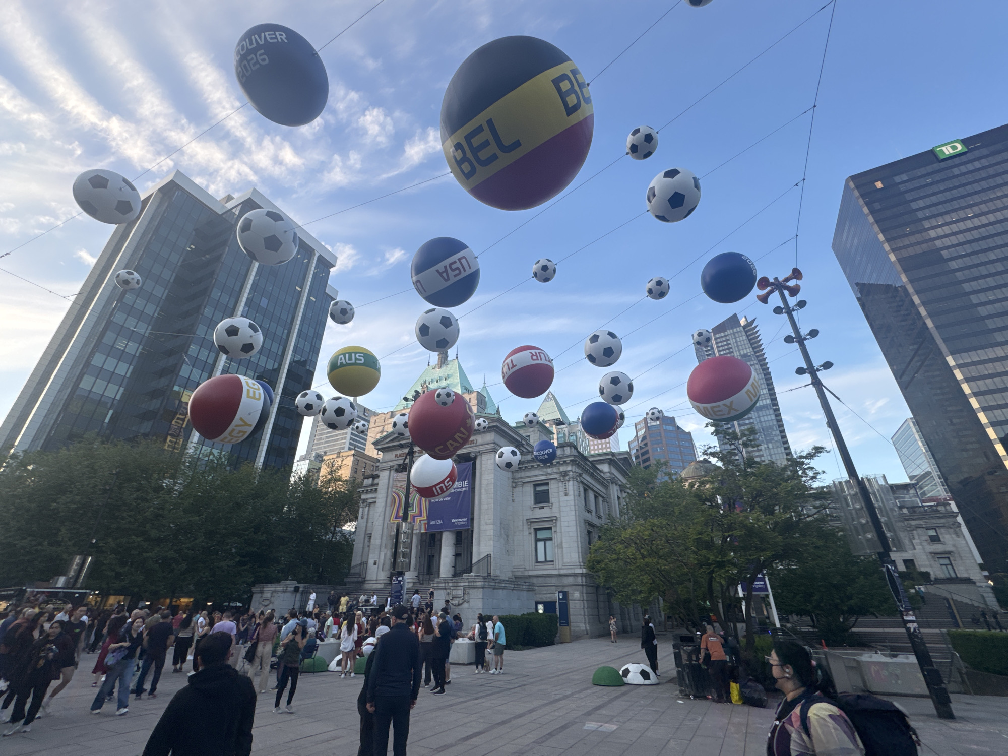 The image depicts a bustling public square in Vancouver, Canada, decorated with overhead soccer-themed decorations. The square is filled with pedestrians, standing, walking, and interacting. In the background stands a historic building adorned with banners, presumably an art gallery, and contemporary skyscrapers flank the scene on both sides. Among the decorations, large spheres painted with national flags like Belgium, Canada, the USA, and others, alternate with standard soccer ball-patterned spheres. A sphere labeled "Vancouver 2026" hangs prominently, hinting at the FIFA World Cup. A TD Bank tower is visible, identifying the city. The setting appears to be sunny and daytime.