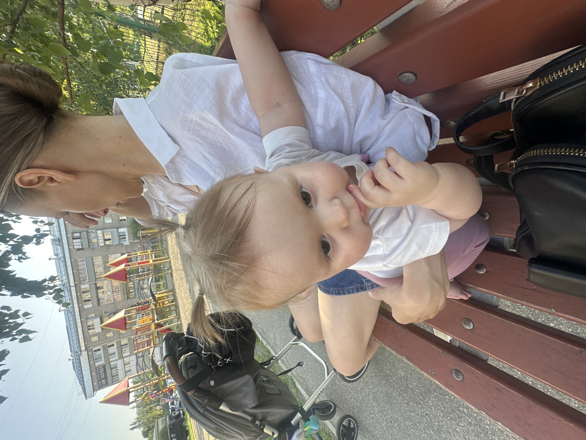 In this vertical, close-up image, a young child lies on a wooden bench, their head angled towards the camera. The child is wearing a white t-shirt and denim shorts, with one hand near their mouth. A woman, partially visible, also sits on the bench, her head turned away from the camera. She wears a white top.
To the right of the bench, a black leather bag with gold zippers is placed. In the background, a playground with colorful structures is visible in front of a multi-story building. The scene appears to be outdoors during the day, with sunlight casting shadows. The overall mood is relaxed and casual, suggesting a family outing in a residential area.
The image is oriented vertically, giving a sense of looking down at the subjects. The background suggests a typical urban or suburban environment in Novoaltaysk, Russia, likely a residential courtyard with a playground. No discernible text is present in the image.