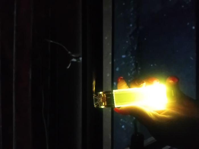 A hand with red painted nails holds a rectangular object, emitting a bright yellow light. The light illuminates the hand and surrounding area. In the background, a darkened scene is visible through a window, possibly showing rain or snow. To the left, a dark curtain partially obscures the view. The object appears to be a light-emitting device, possibly a lighter or a small lamp, casting a strong glow.