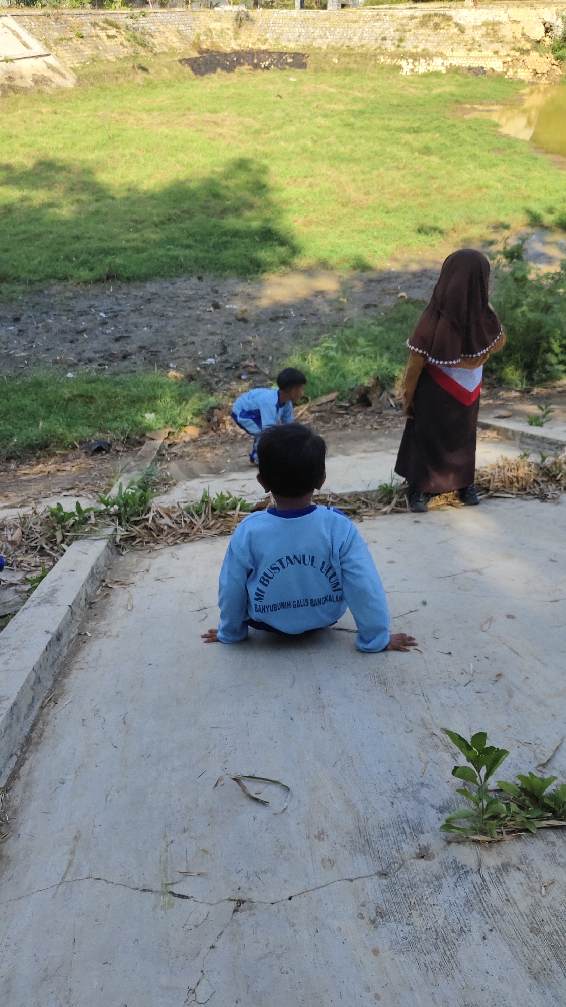 In Banyubunih, Indonesia, three children are observed in an outdoor setting with a grassy embankment and a body of water. A young child, facing away from the camera and seated on a concrete surface, wears a light blue long-sleeved shirt with writing on the back that appears to say "MI BUSTANUL ULUM BANYUBUNIH GALIS BANGKALAN". Another child in a similar light blue outfit is crouched down further away, facing the grassy area. A third child, wearing a brown hijab and a tan outfit with a red and white sash, stands near the grassy embankment, also facing away from the camera. The environment includes dry leaves scattered on the ground, sparse vegetation, and a cracked concrete path. The overall scene suggests a casual outdoor activity, possibly near a school or communal area, given the writing on the child's shirt.