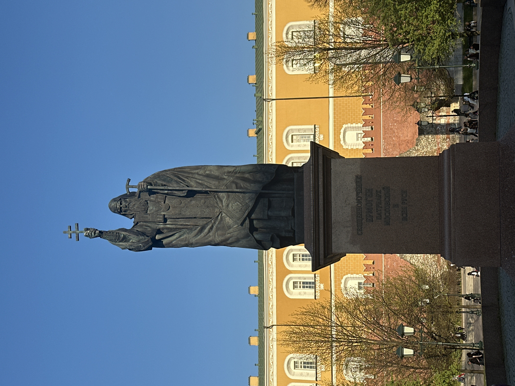 The image depicts the statue of Patriarch Hermogenes in Moscow, Russia, set against a clear blue sky. The monument stands on a stone pedestal with Cyrillic inscriptions. In the background, there's a section of the Moscow Kremlin wall with its distinctive architectural details, including brickwork and arched windows. People are visible walking along the sidewalk and sitting on benches near the wall. Street lamps are spaced along the walkway, interspersed among the trees.