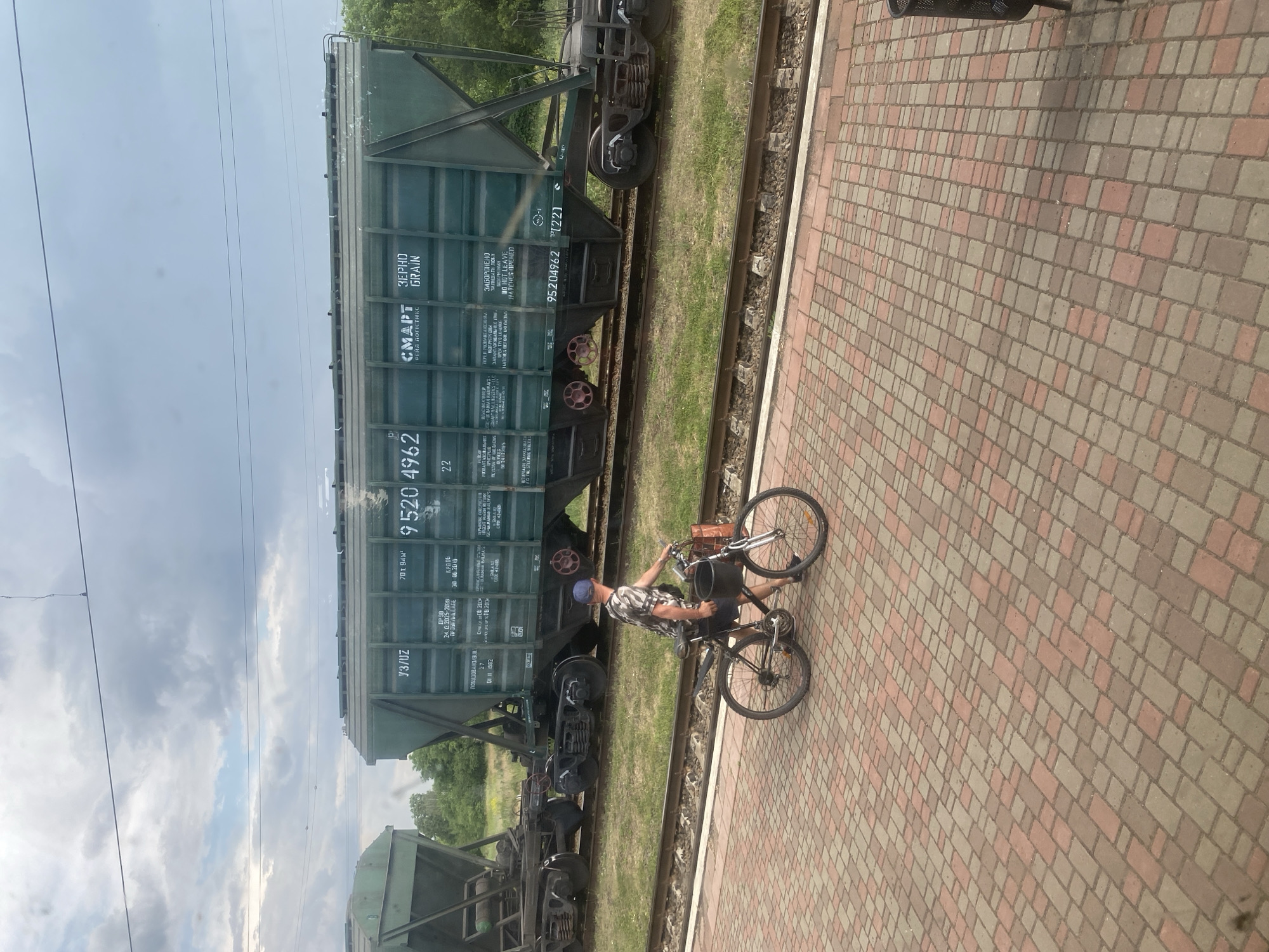 A man wearing a camouflage t-shirt and dark shorts, with a blue cap on his head, is walking a bicycle on a brick-paved platform. The bicycle has a basket on the front and a bag on the rear rack. Behind the man, a long, green train car with white lettering is parked on railway tracks. The train car is a hopper car designed for transporting grain, as indicated by the text "ЗЕРНО GRAIN" on its side. The environment appears to be a train station in Myrhorod, Ukraine, with grassy areas and trees visible beyond the train tracks under a cloudy sky.