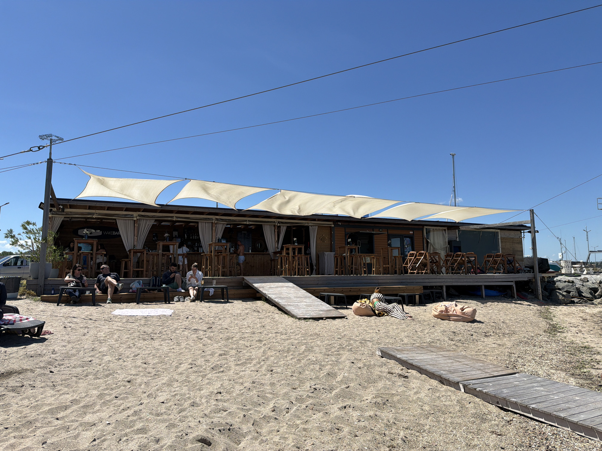 The image shows a beach bar in Burgas, Bulgaria on a sunny day. The bar is a single-story wooden structure with a series of white shade sails above it. There are people seated at tables on a deck in front of the bar, as well as on benches closer to the sand. A wooden ramp provides access from the deck to the beach. A wooden walkway extends across the sand toward the right side of the frame, where there are rocks, sailboats and a dock. Powerlines run above the building. The beach is sandy and slopes up towards the bar. Several people are relaxing on the beach on blankets and bean bag chairs. A silver vehicle is parked behind the bar on the left side. The sky is clear and blue.