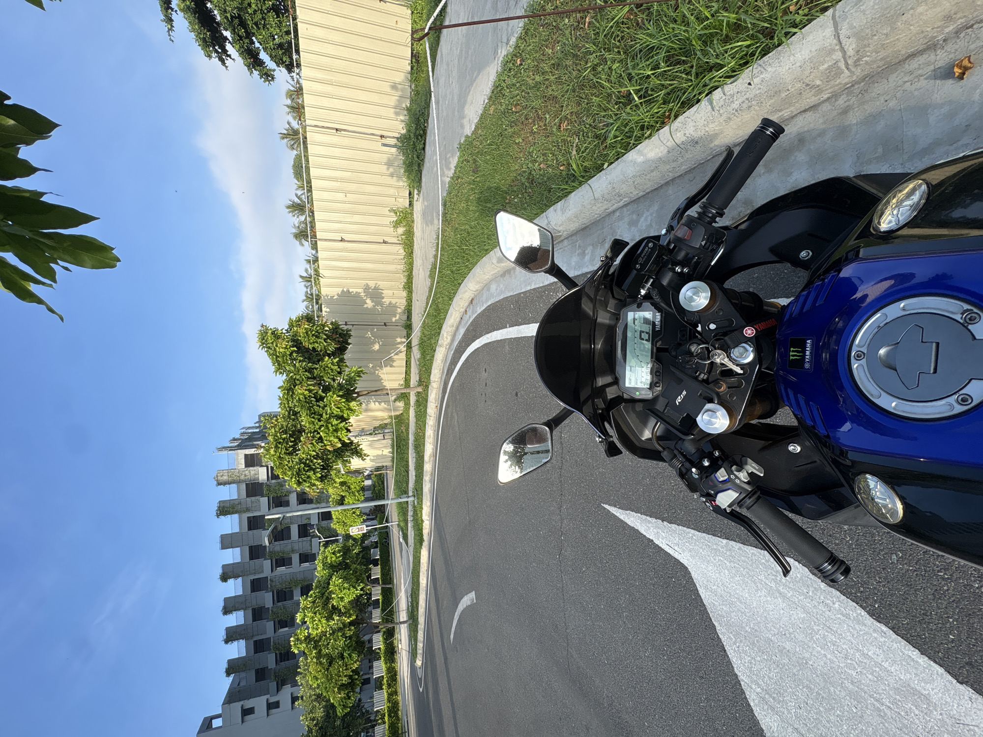 The image shows a street scene in Davao City, Philippines, on a clear day with a blue sky and some clouds. A blue and black Yamaha motorcycle is parked on the right side of the road, with its front facing forward. The motorcycle has a digital display on the dashboard, showing "0.0" and "231584". The road has a white lane marking that curves to the left. To the right of the road, there is a concrete curb and a strip of green grass, followed by a light-colored metal fence. In the background, there is a partially constructed building with a modern architectural design, lined with trees. Further in the distance, palm trees are visible. The lighting suggests it is either early morning or late afternoon.