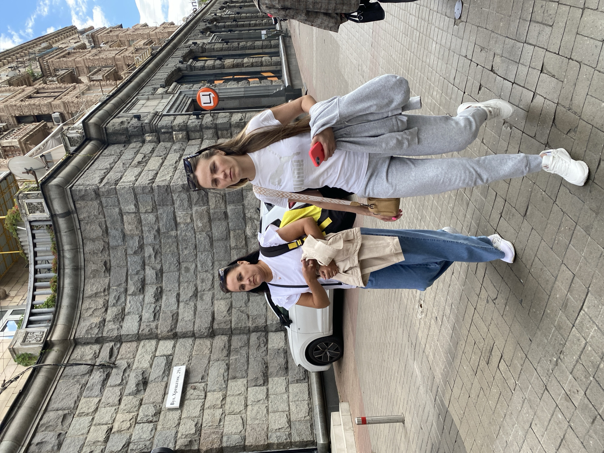 Two women walk on a paved city sidewalk in front of a stone building. The woman on the left, wearing blue jeans, a white t-shirt, and white sneakers, carries a beige jacket over her arm and a yellow bag. The woman on the right, wearing grey sweatpants, a white t-shirt with a Puma logo, and white sneakers, holds a red phone and a grey jacket. They are walking towards the viewer. A white car is parked on the street behind them. The building behind them is made of large stone blocks and has ornate architectural details. A street sign reads "вул. Хрещатик, 29". The sky is partly cloudy.