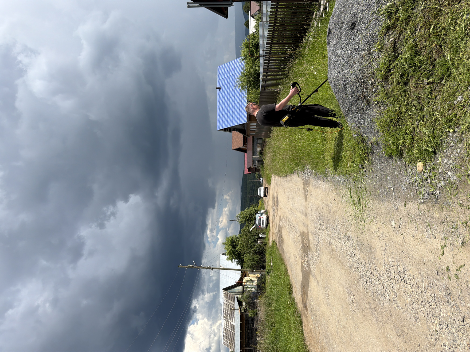 The image depicts a rural scene in the city of Mezhevoy, Russia, under a dramatically cloudy sky. A man is using a string trimmer to cut grass along a dirt road, next to a pile of gravel. The road curves to the left, bordered by grass and vegetation. Several houses with wooden fences are visible on the right side of the road, one of which has a bright blue metal roof. On the left side of the road, there are more houses and trees. A few cars are parked or driving on the road in the distance. The overall atmosphere suggests preparation or outdoor maintenance in a village setting, possibly anticipating rain due to the dark clouds.