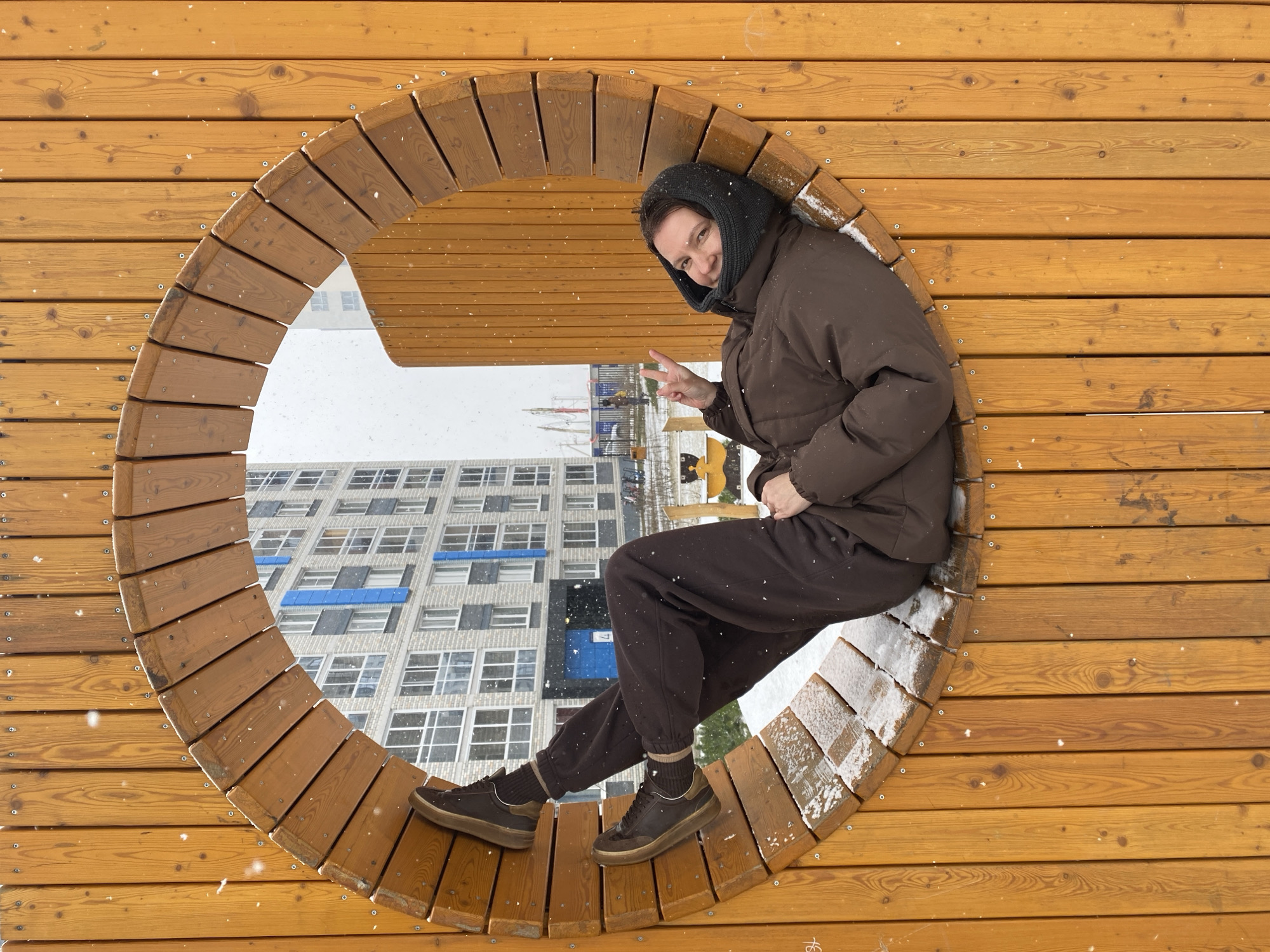 The image shows a person sitting in a wooden circular structure, with a view of a building and snowy weather visible through the opening. The person is wearing a brown jacket, sweatpants, and brown shoes and a dark headscarf, and they are giving a peace sign. The circular structure appears to be a part of a playground or outdoor installation. The scene is likely in Yekaterinburg, Russia, judging by the style of architecture visible in the background. It is snowing.