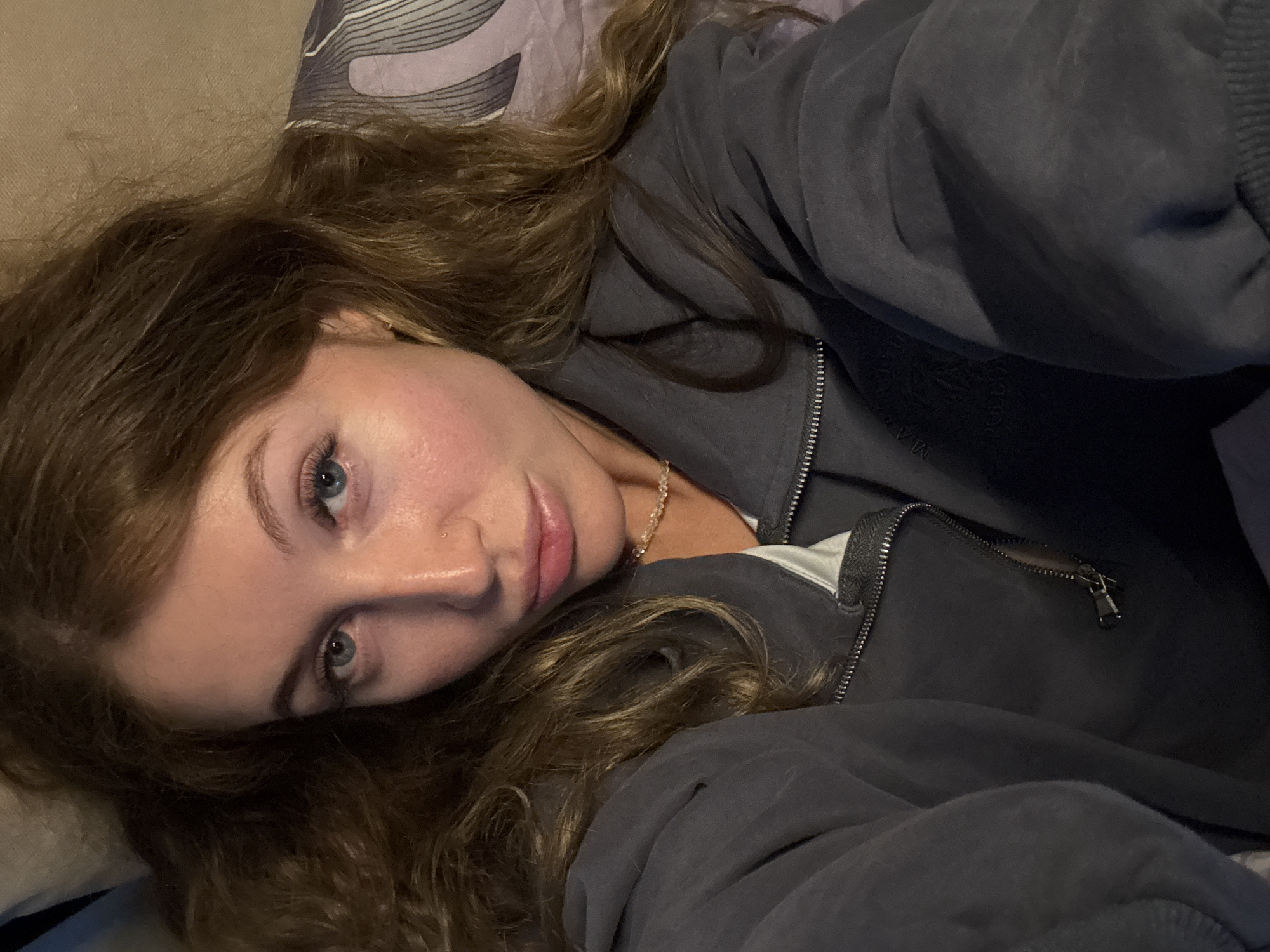 A young woman with fair skin and wavy brown hair is lying down. She is wearing a dark gray zip-up jacket and a necklace. Her eyes are blue. The background includes bedding with a patterned pillow. There is no discernible information to place the location in the city of Chaikovsky, Russia.