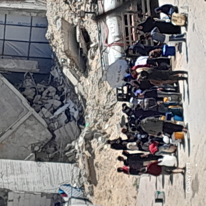 This image depicts a scene of civilian activity amidst extensive urban destruction in Khan Yunis, Palestinian Territory. In the background, a large building is reduced to rubble, with concrete slabs, twisted rebar, and debris forming a significant mound. Structural elements of partially standing buildings are visible, showing shattered windows and exposed interiors. In the midground, a group of approximately 30-40 individuals, comprising men, women, and children, are gathered. Many individuals are carrying or standing near buckets, jerrycans, and bags. To the right, two large, white horizontal tanks are visible, appearing to be water storage or distribution tanks, resting on wooden pallets. One tank has discernible Arabic script on its side. The ground beneath the people is dusty and uneven. The overall scene shows a concentration of people seemingly collecting or waiting for essential supplies in a severely damaged environment.