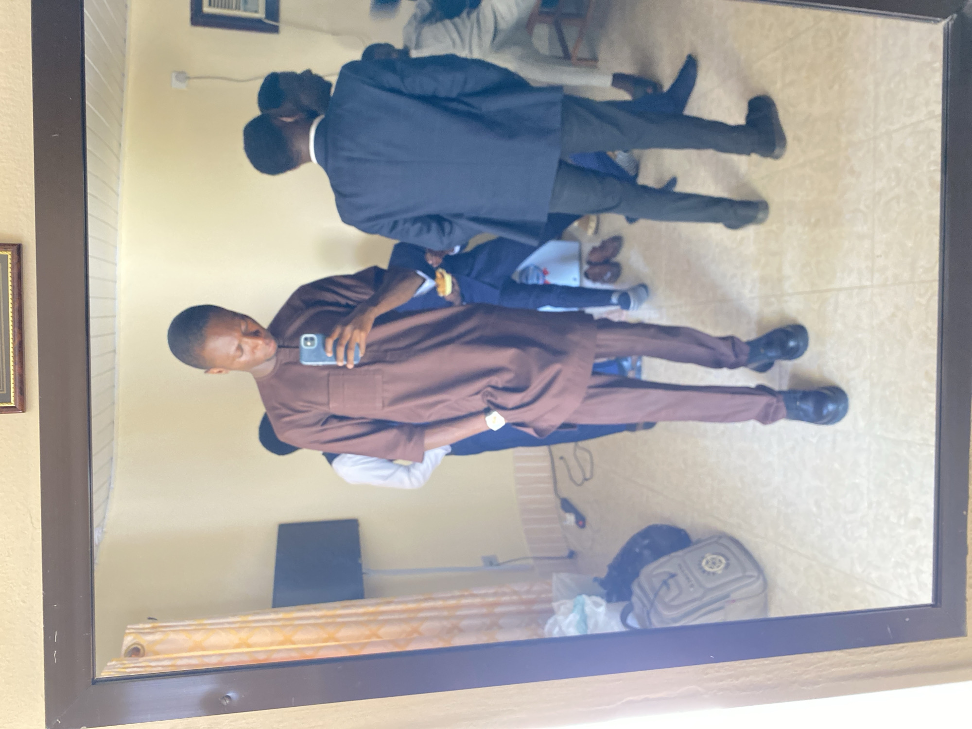 A mirror reflects a scene in what appears to be an interior space in Barakin Angundi, Nigeria. A man is holding a phone, seemingly taking a picture or a selfie, wearing a long-sleeved brown outfit and black shoes. Behind him are several other people. One man is wearing a suit. A television is visible on the wall. There is a bag on the floor and a curtain.