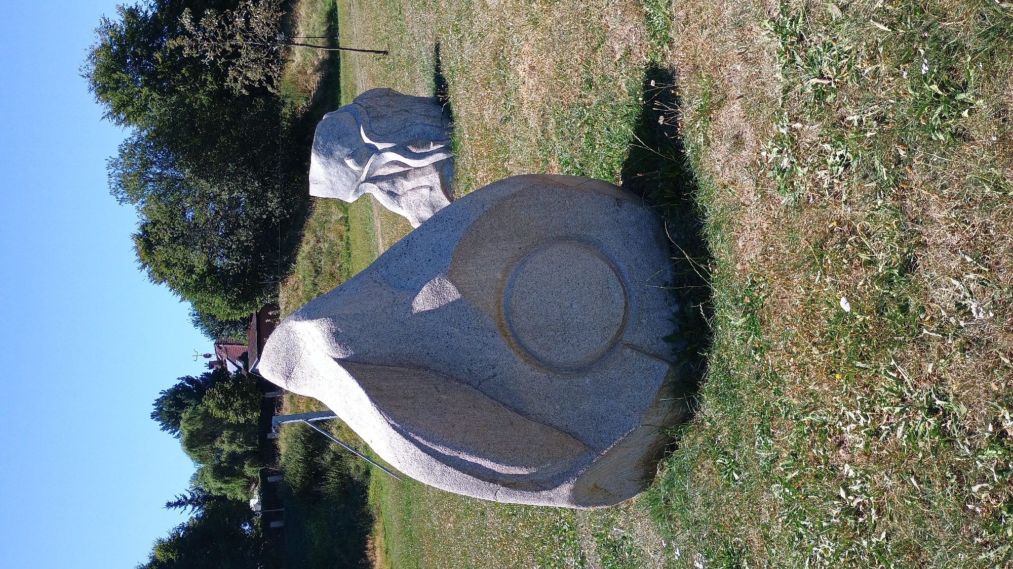 This is a photograph taken outdoors in Sokolov, Czechia. The scene features two large, abstract stone sculptures in a grassy area. The sculpture in the foreground is a large, grey, roughly egg-shaped rock with carved indentations. The second sculpture, located behind and to the right of the first, is also made of grey stone and appears more fluid and organic in form. The background shows a line of trees and a few buildings, suggesting a semi-rural or park-like setting within the city. The sky is clear and blue. The ground is covered in dry grass with some patches of green. There are no people visible in the image.