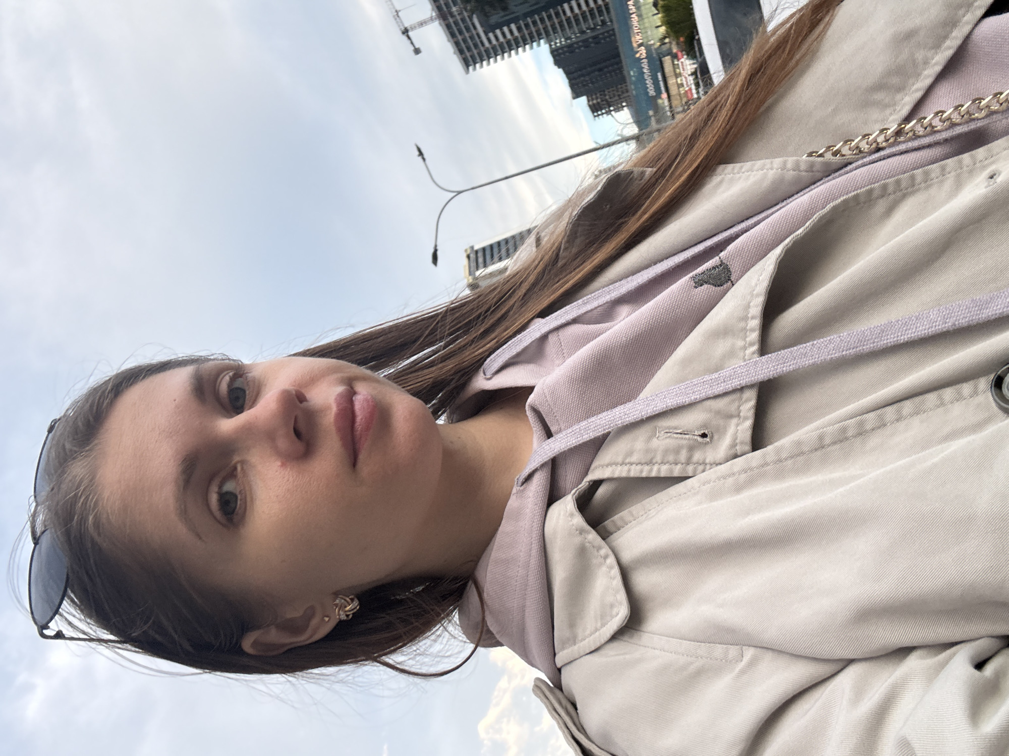 A young woman is posing for a photo in Batumi, Georgia. She has light brown hair and is wearing a light beige trench coat over a lavender hoodie. Sunglasses are perched on top of her head, and she has small, intricately designed gold earrings. The background features modern architecture, including tall buildings, one of which is under construction with a crane visible. The sky is overcast, and a street lamp is partially visible. The photo is taken at a slightly tilted angle.