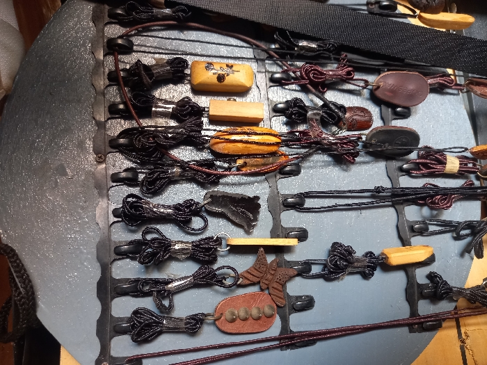 The image shows a display of various handcrafted items hanging on a metal rack, likely for sale. The rack is mounted on a wall painted a light shade of blue, with a dark brown wooden frame surrounding the display. The rack is made of metal bars with hooks along its length.
Various items, including pendants, necklaces, and charms, are hanging from the hooks. The pendants, primarily crafted from wood and featuring carvings, are in shades of brown and dark brown. Some have intricate designs, while others are plain. The majority of the hanging items are attached to black cords, but some are secured with brown or black leather straps.
The image suggests a local market or craft shop in Ita Oniyan, Nigeria. The items likely represent traditional craftsmanship and are potentially sold to locals or tourists. The precise location cannot be determined from the image alone, but it depicts an interior space with the potential for a storefront.