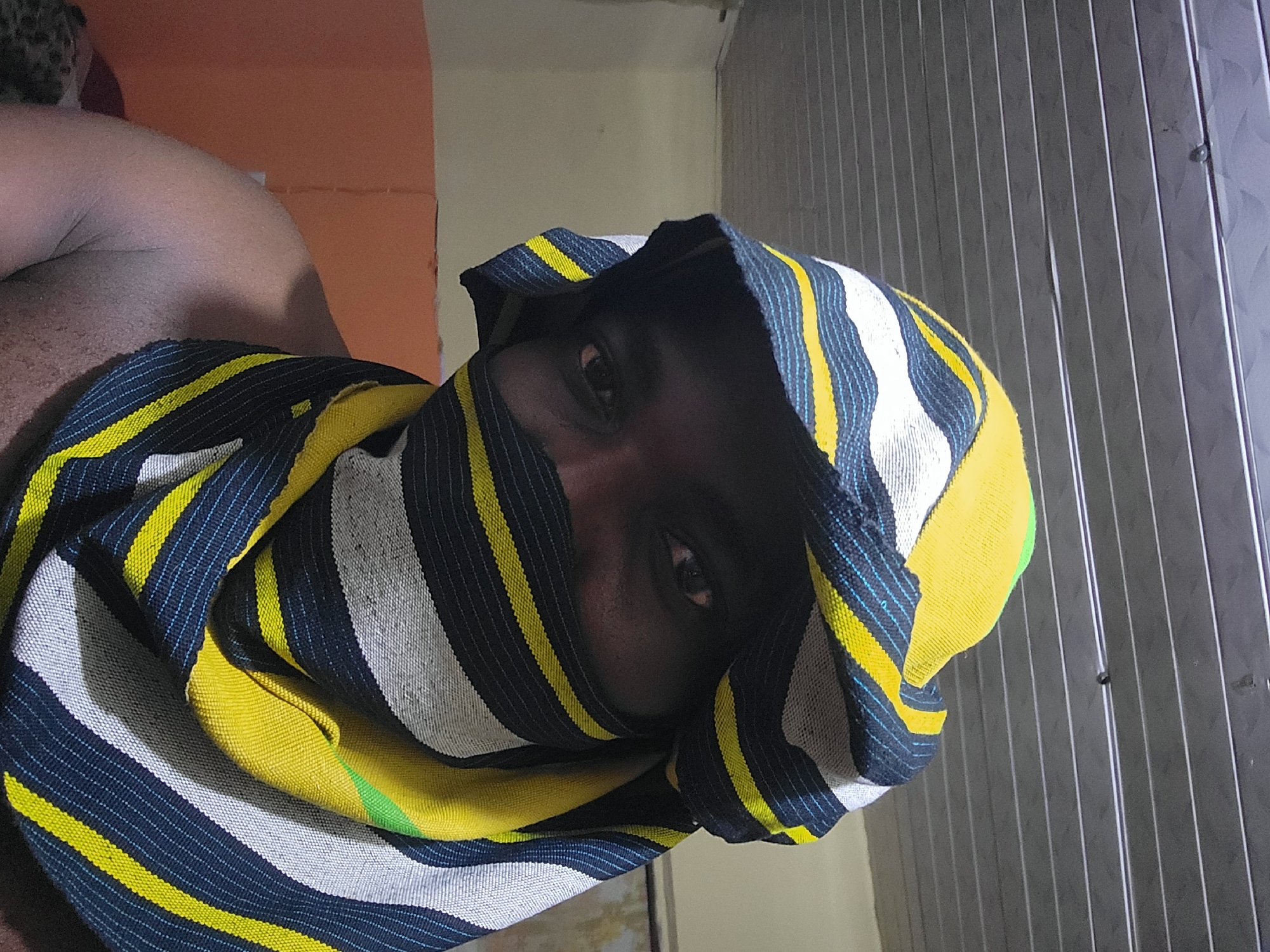 This indoor image from the city of Jos, Nigeria, features an individual looking directly into the camera. The person has dark skin, with only the eyes and upper facial area visible; the remainder of the face is covered by a patterned textile wrap. The wrap displays prominent horizontal stripes in dark blue, white, and bright yellow, incorporating thin light blue filaments within the dark stripes and a small vibrant green section on a yellow stripe. The fabric forms a hood-like covering over the head and extends around the neck and lower face. A bare shoulder and upper arm of the individual are visible on the left side of the frame. The background consists of a gray, horizontally paneled ceiling, likely corrugated metal or PVC, with visible fixtures. Behind the individual, a light-colored wall (possibly beige) is partially visible on the left, with an orange-brown surface below it.