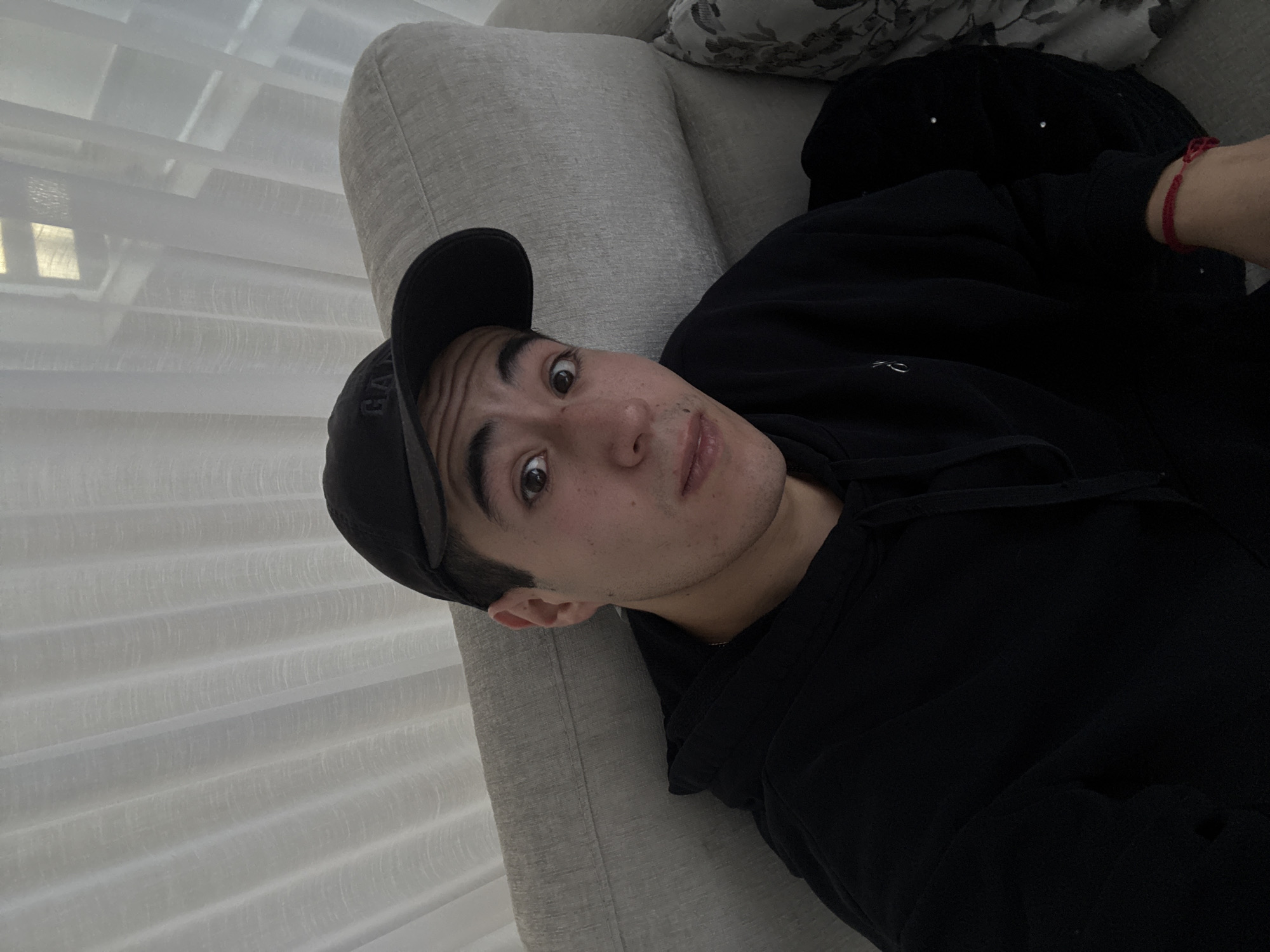 A young man is depicted lying on a light grey sofa indoors, captured from a slightly elevated angle. He has dark hair, dark eyes, and a light to medium skin tone, looking directly at the camera with wide-open eyes and slightly parted lips, conveying a surprised or curious expression. He wears a black baseball cap with the letters "GAP" faintly visible on the front, and a black hooded sweatshirt. A red string bracelet adorns his right wrist. Behind his head, a patterned cushion rests on the sofa. To the left of the frame, a window covered by white horizontal blinds or sheer curtains allows soft, diffused daylight to illuminate the scene. The overall setting suggests a casual, relaxed indoor environment. The image is rotated approximately 90 degrees clockwise. No other specific location cues are visible within the frame.