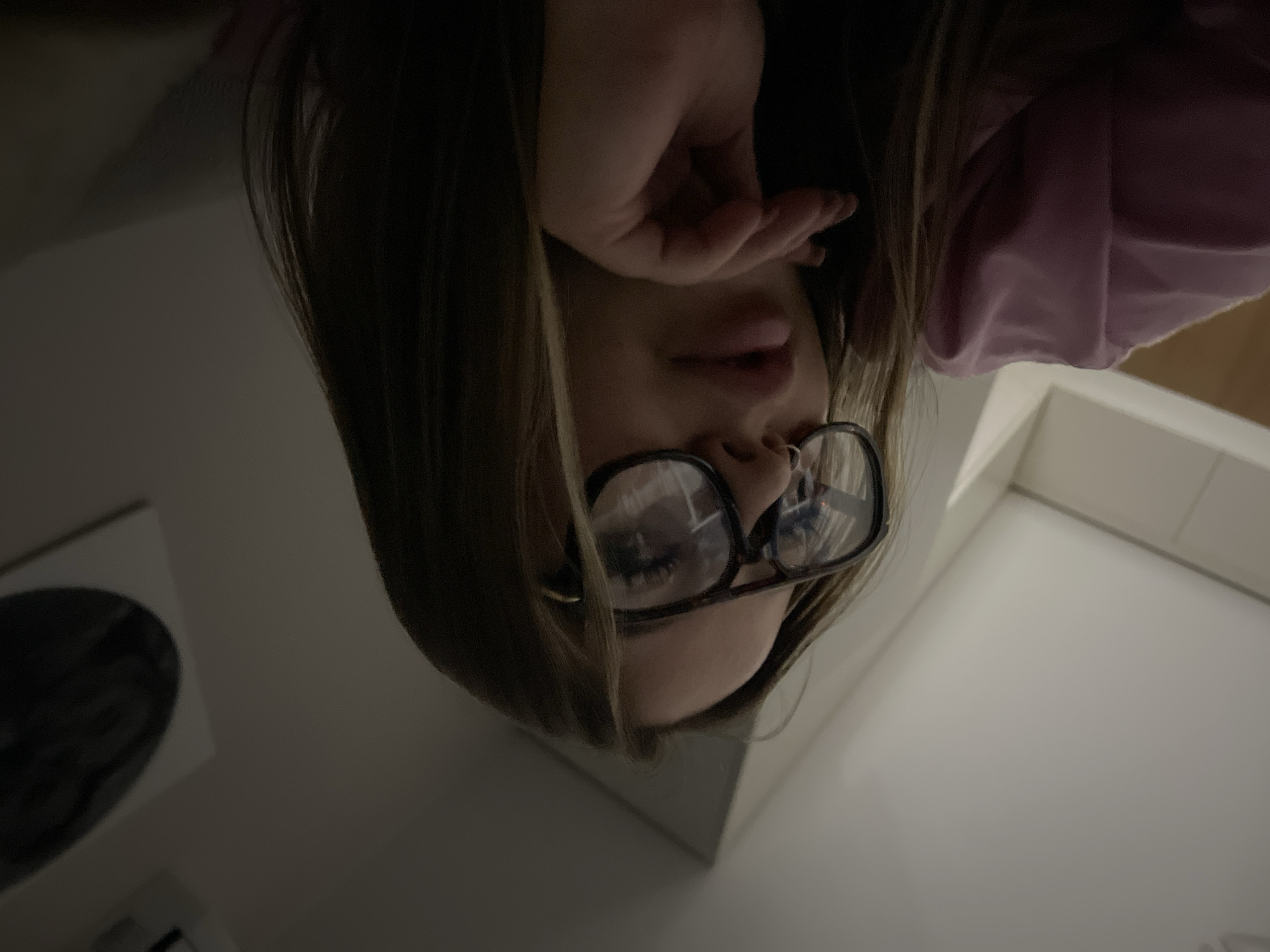 A woman with fair skin, wearing glasses and a pink top, is shown from an upside-down perspective. Her hand is partially covering her face. The background includes a white surface, possibly a countertop or wall, and a framed object on the left. The location cannot be determined.
