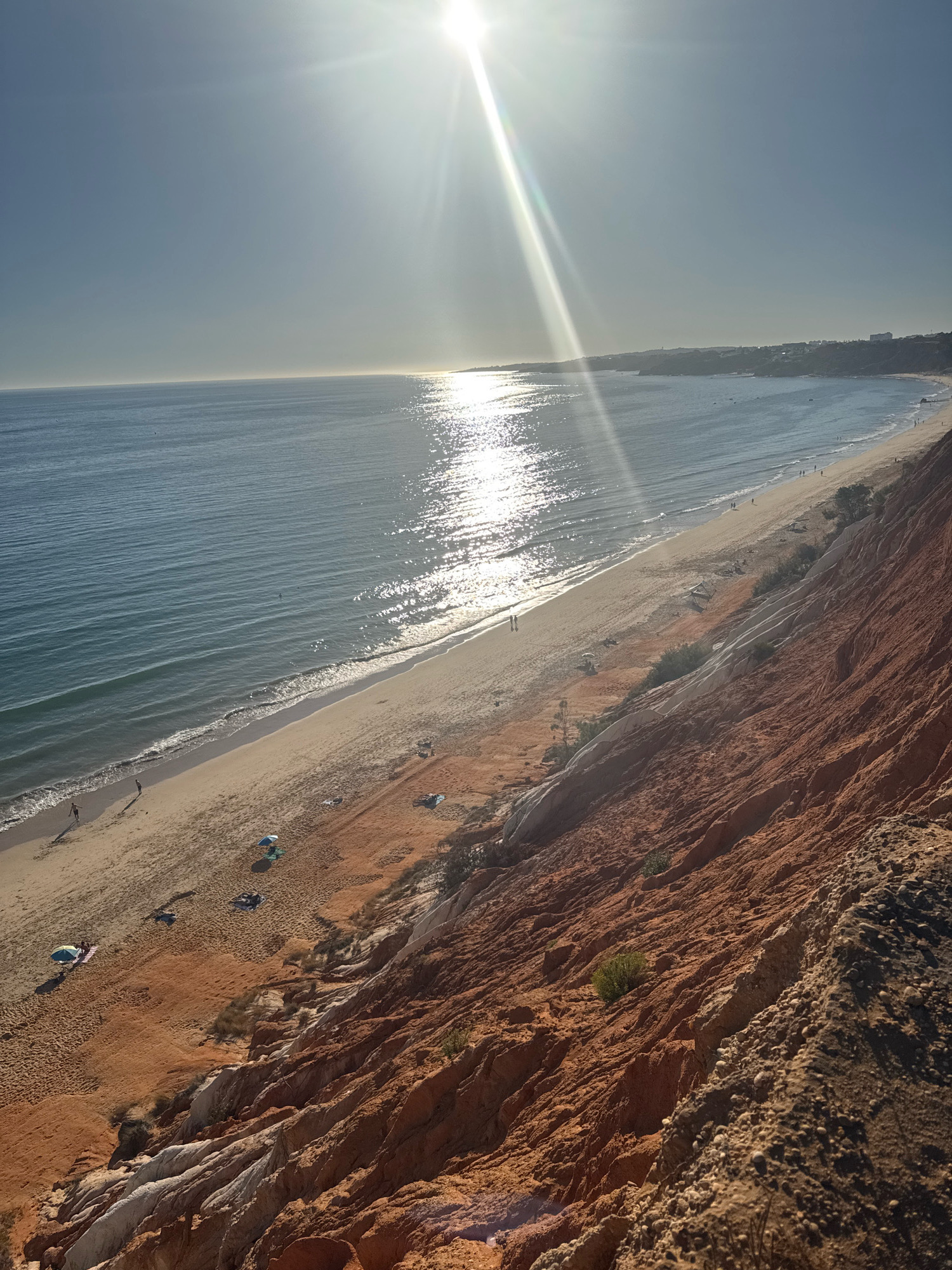 The image depicts a wide, scenic view of a beach in Albufeira, Portugal, under bright sunlight. The sea, a tranquil blue, stretches to the horizon, reflecting the sun's glare. Along the coastline, a long, sandy beach is visible, dotted with a few beach umbrellas, towels, and several individuals. Some people are seen walking along the shore, while others are relaxing on the sand. The beach is bordered by an impressive, multi-colored cliff face, characterized by layers of red, orange, and white sedimentary rock. Sparse vegetation grows along the slopes of the cliff. In the distance, a town or resort area can be seen nestled along the top of the cliffs. The overall scene conveys a sense of a peaceful, sunny day at a popular coastal destination.